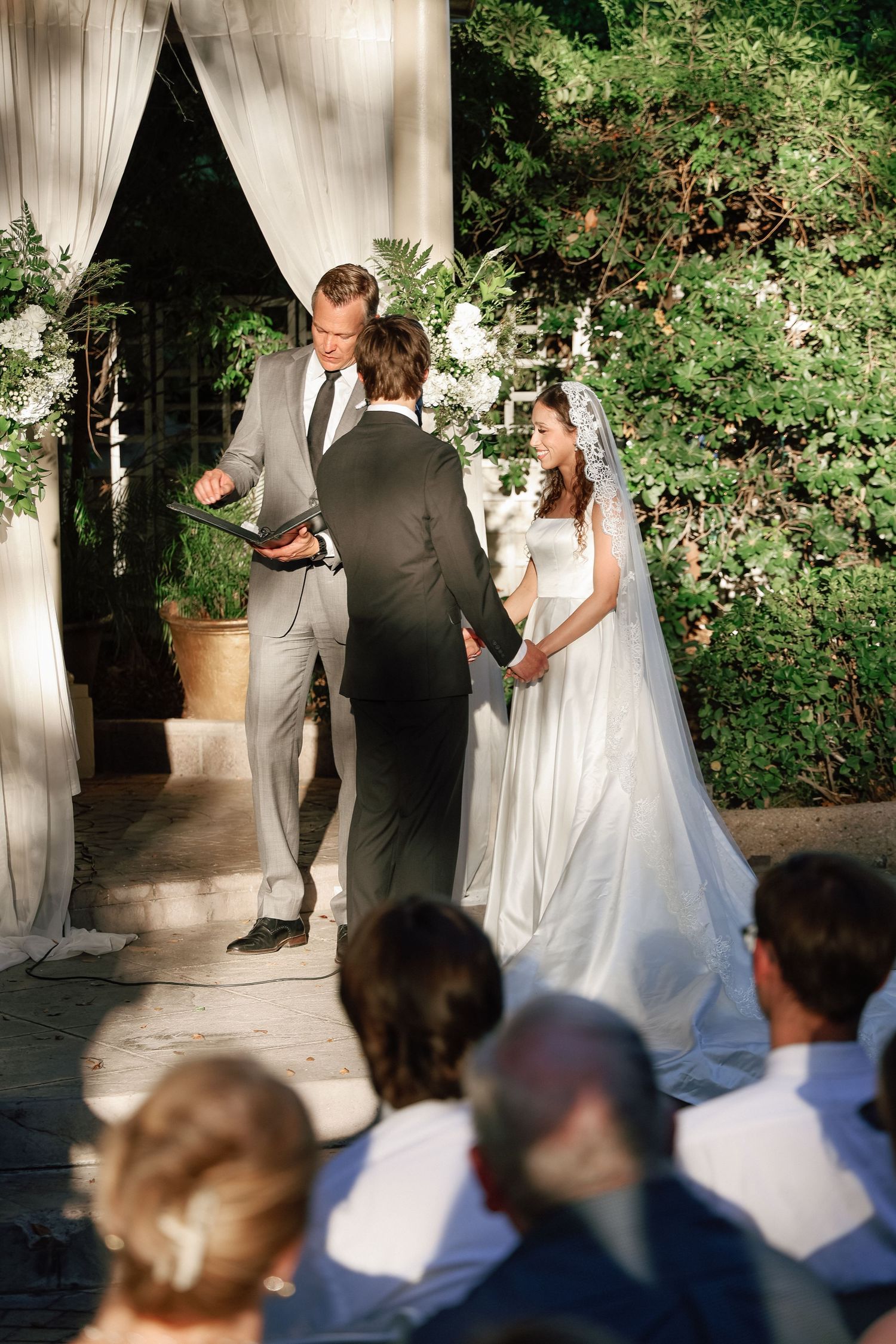 Bride and groom exchange vows at an elegant outdoor wedding ceremony with white floral arch and seated guests.
