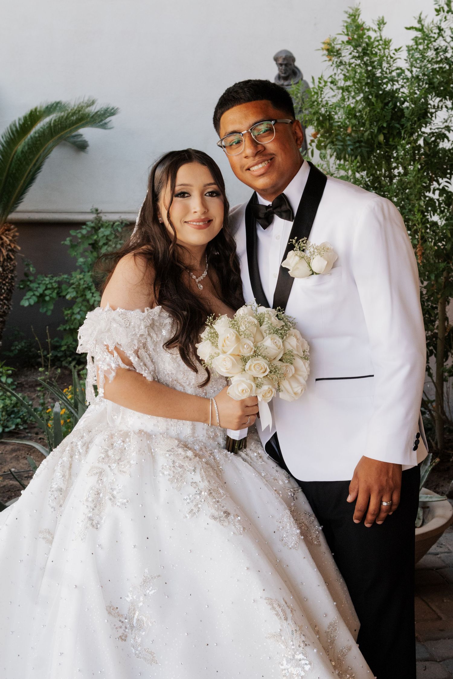 A wedding couple poses together, bride holding white rose bouquet and groom in white dinner jacket.