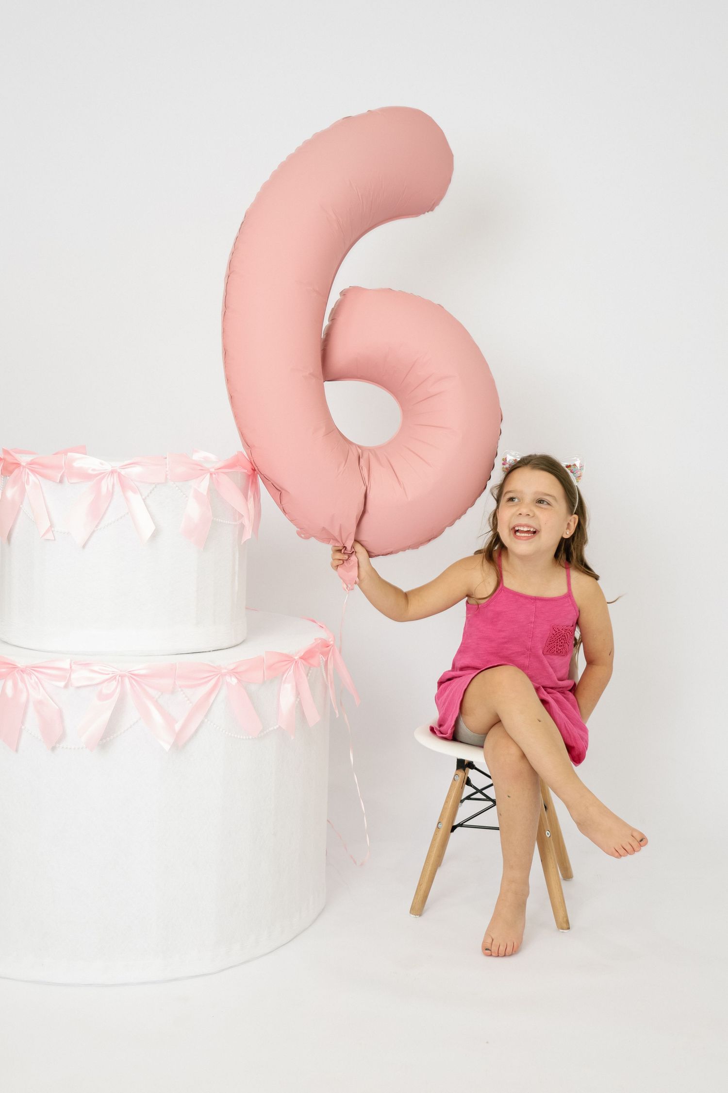 A happy girl in pink dress sits beside a white cake, holding a pink number 6 balloon on her 6th birthday.