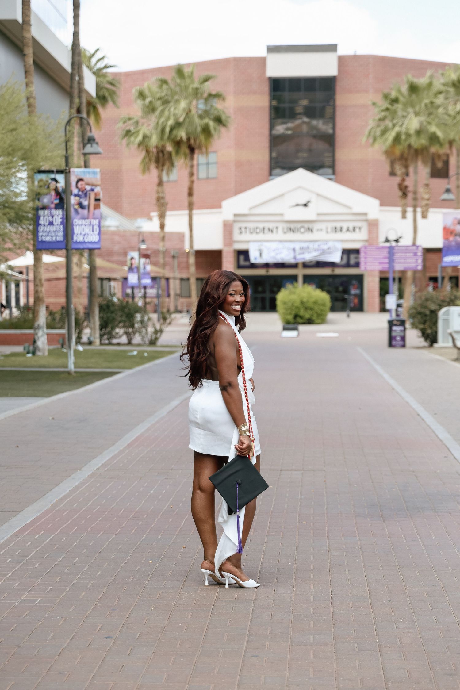 A woman in a white dress and heels poses on a brick plaza outside a university Student Union and Library building.