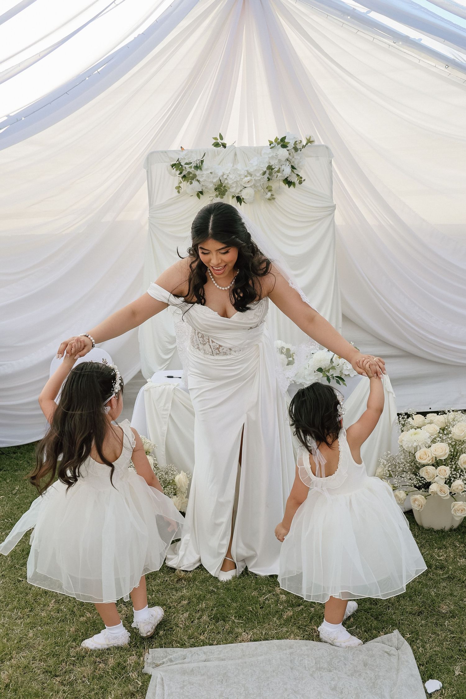 Bride in white gown holds hands with two flower girls in white dresses under draped fabric canopy at outdoor wedding.