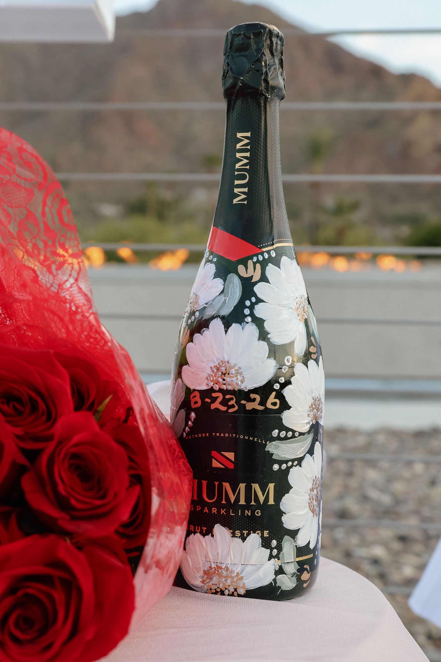 Mumm Sparkling champagne bottle with floral design and date 8-23-26 beside red roses on white table.