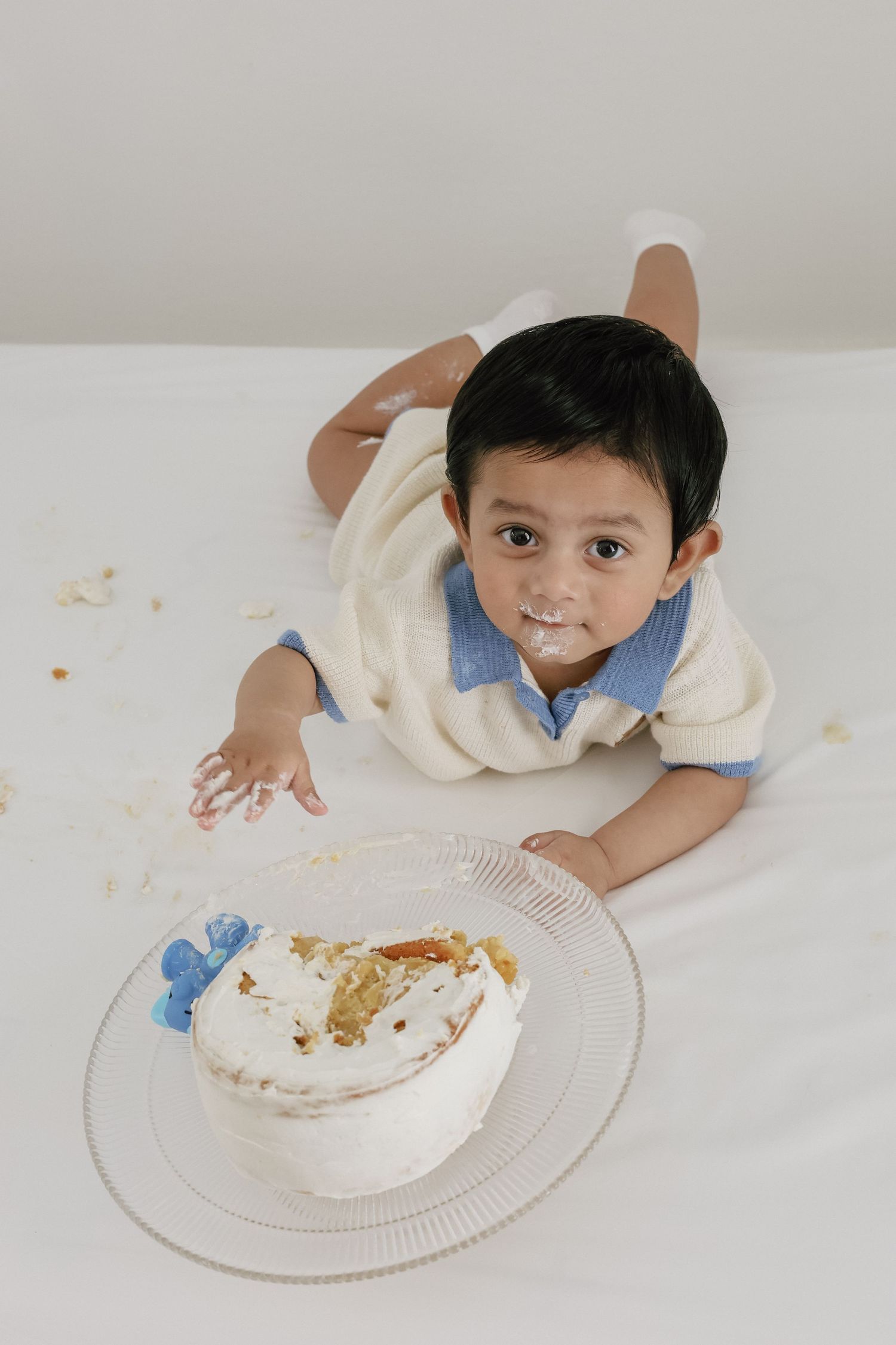 Toddler boy with cake on his face lying next to a smashed white birthday cake on a white surface.
