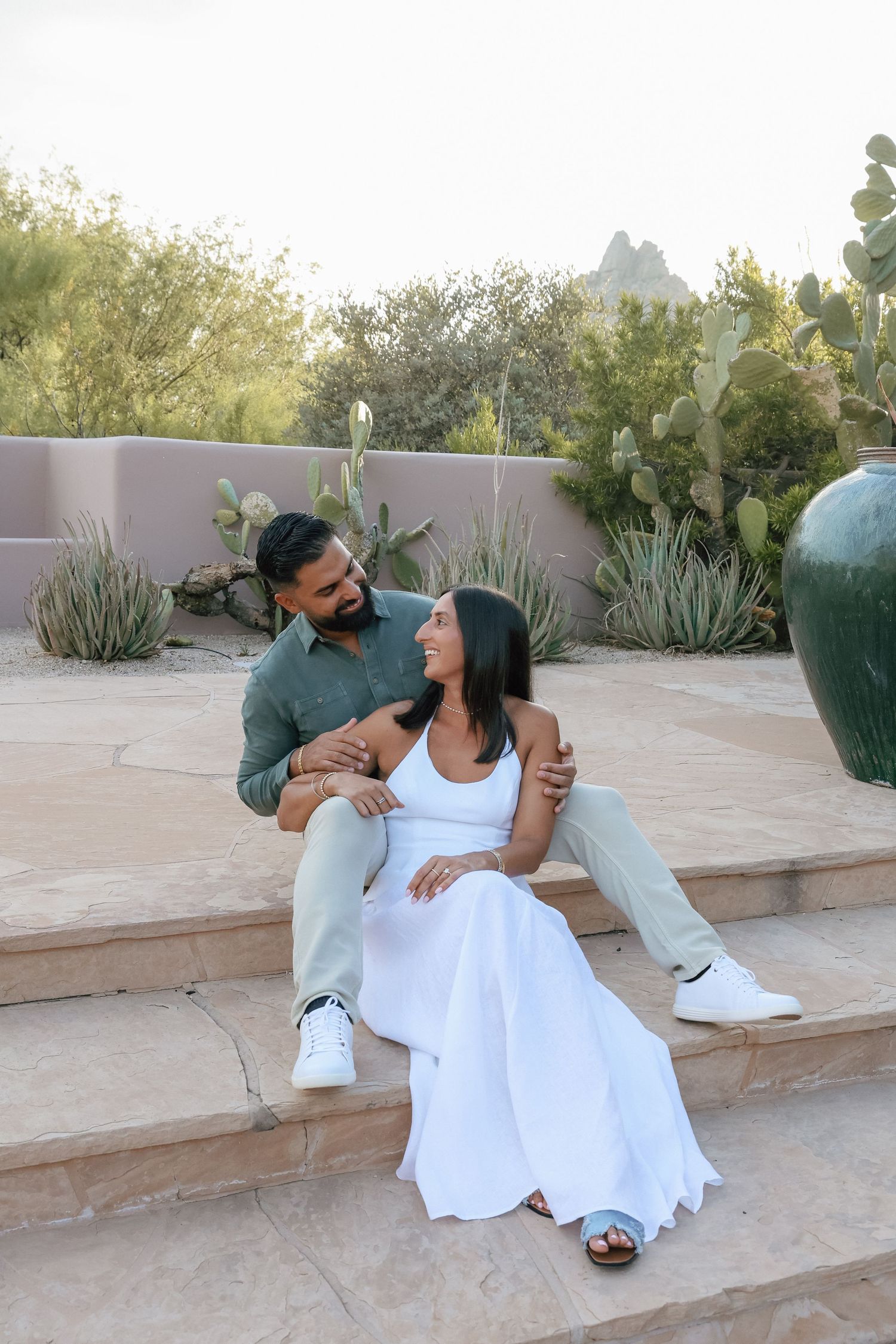Couple sitting on stone steps in a desert garden, woman in white dress leaning on man in green shirt.