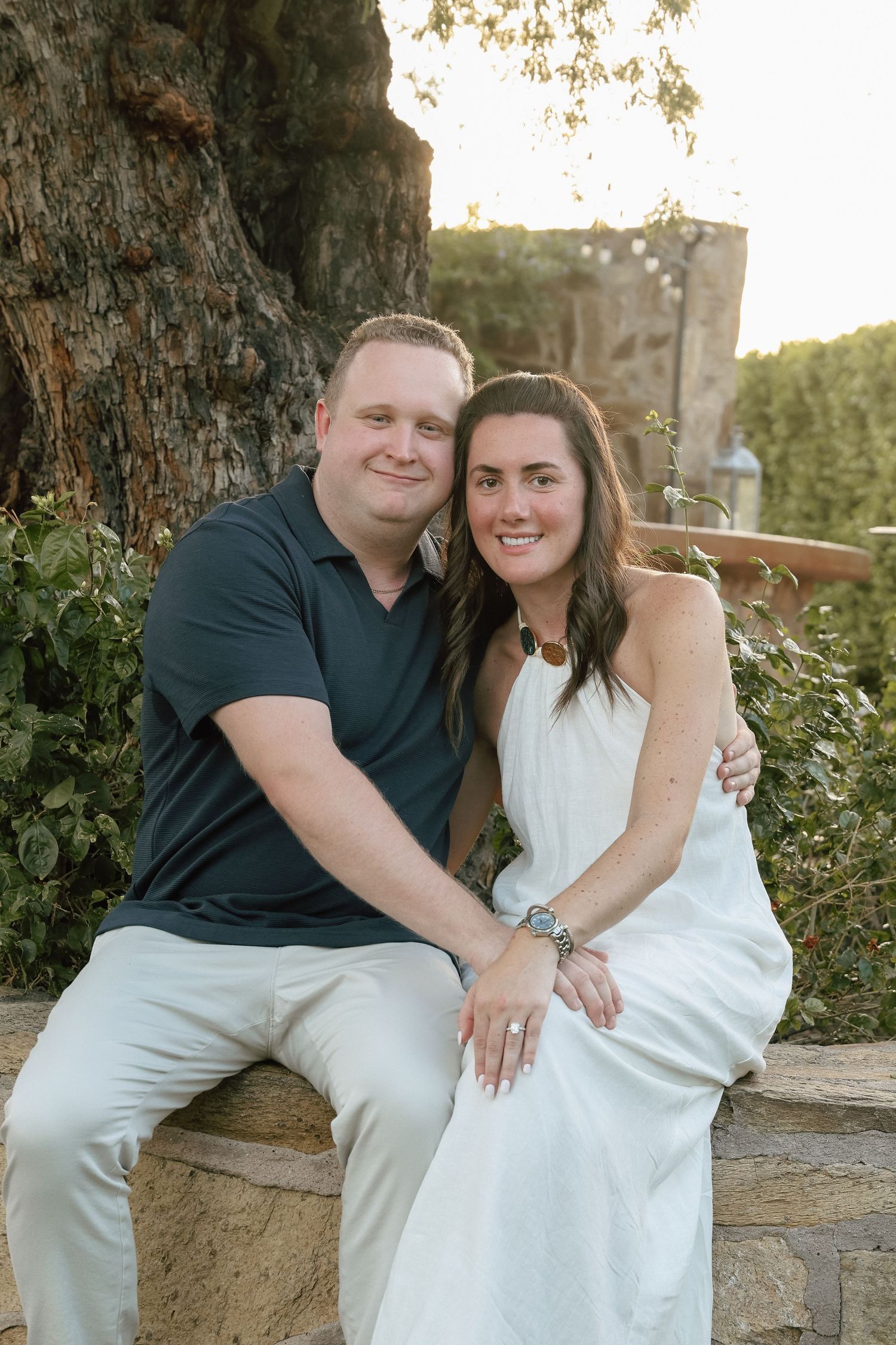 A smiling couple poses outdoors on a stone wall, man in navy polo and woman in white dress, surrounded by lush greenery.