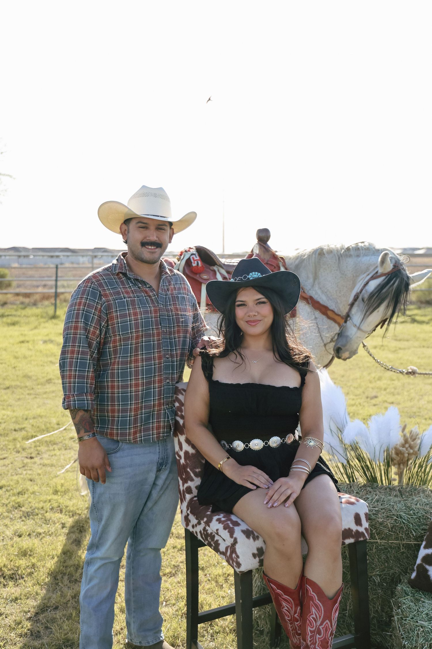 A couple in cowboy hats pose outdoors near a white horse and hay bale at a western-themed event.