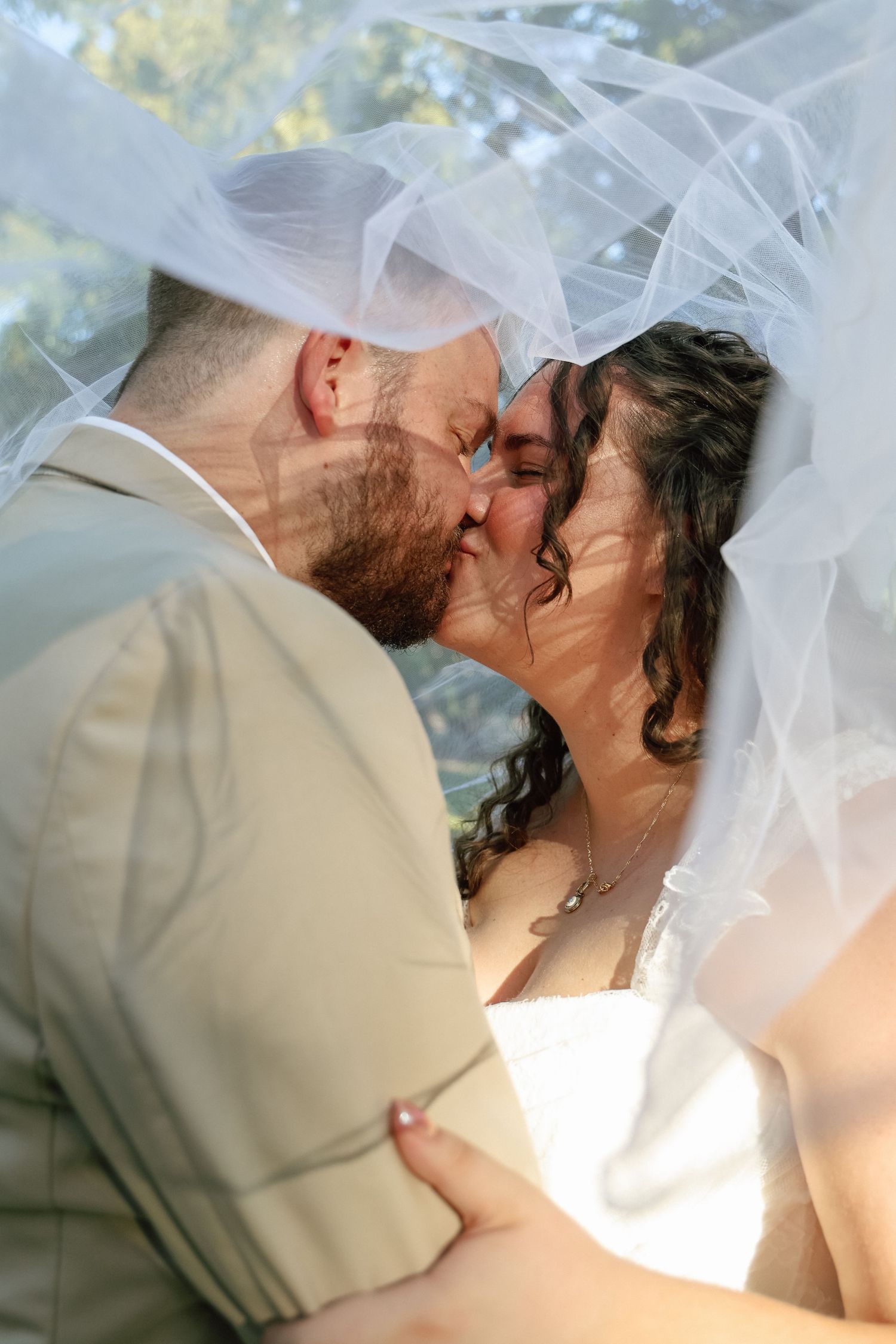 Bride and groom share a romantic kiss under a flowing white veil outdoors on their wedding day.