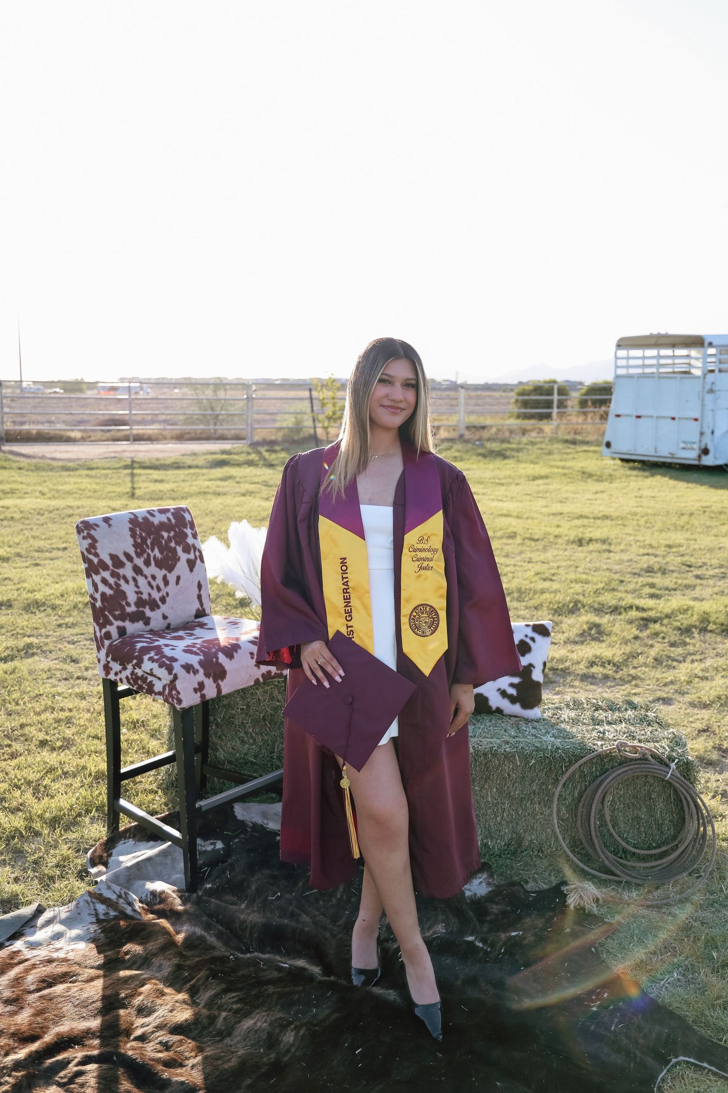 Young woman in maroon graduation gown and gold honor stole poses on hay bale outdoors at golden hour.