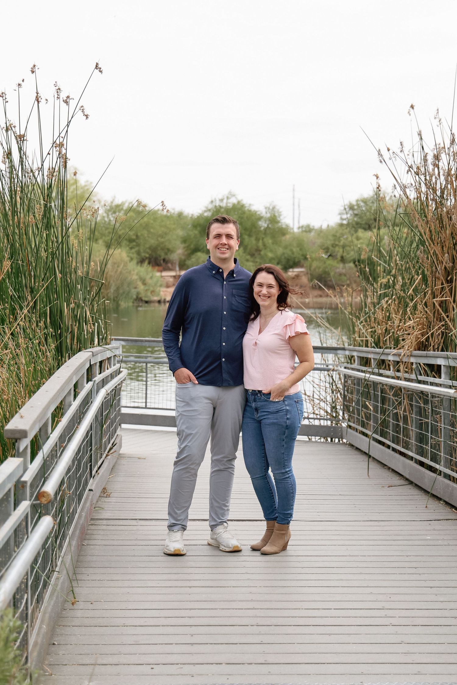 A smiling couple poses together on a wooden boardwalk bridge surrounded by reeds and a lake in a nature park.