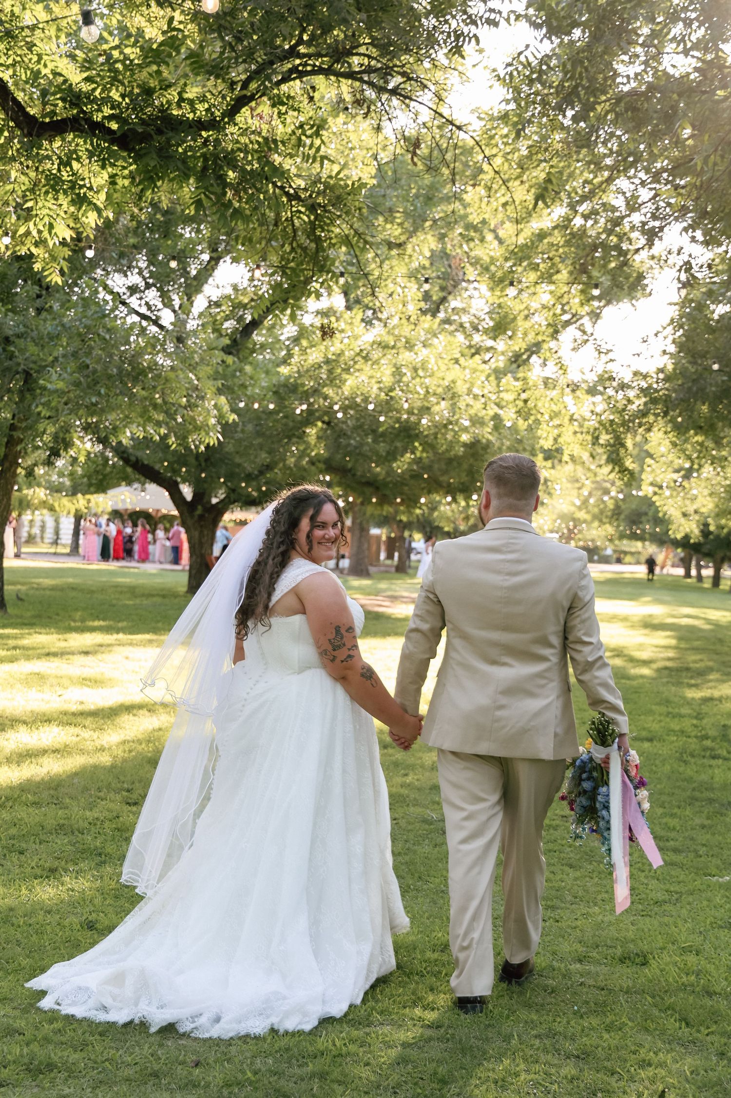 Bride and groom walking hand in hand through a sunlit green park, bride wearing a white gown with veil.
