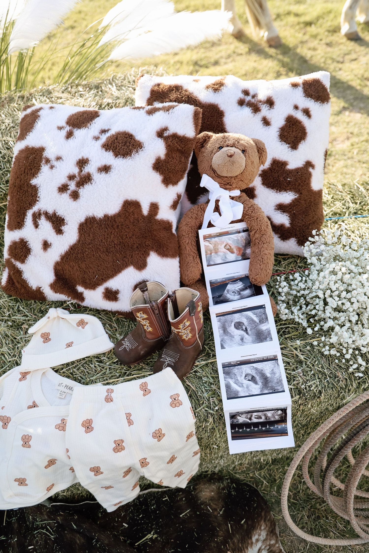 Cow print pillows, teddy bear, ultrasound photos, baby clothes, and cowboy boots arranged on grass for a western baby shower.