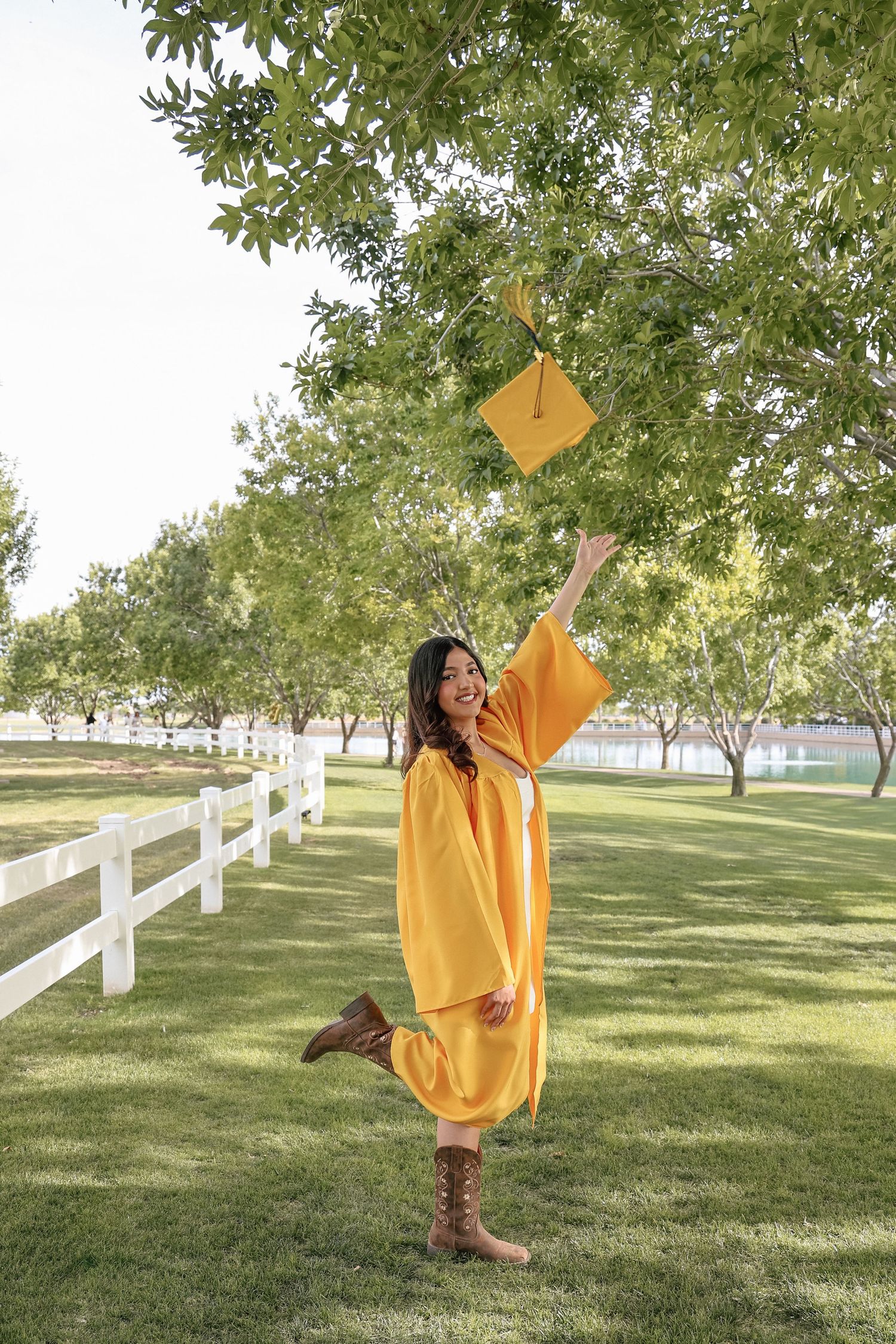 Young woman in golden graduation gown tossing cap in the air outdoors, celebrating academic achievement.
