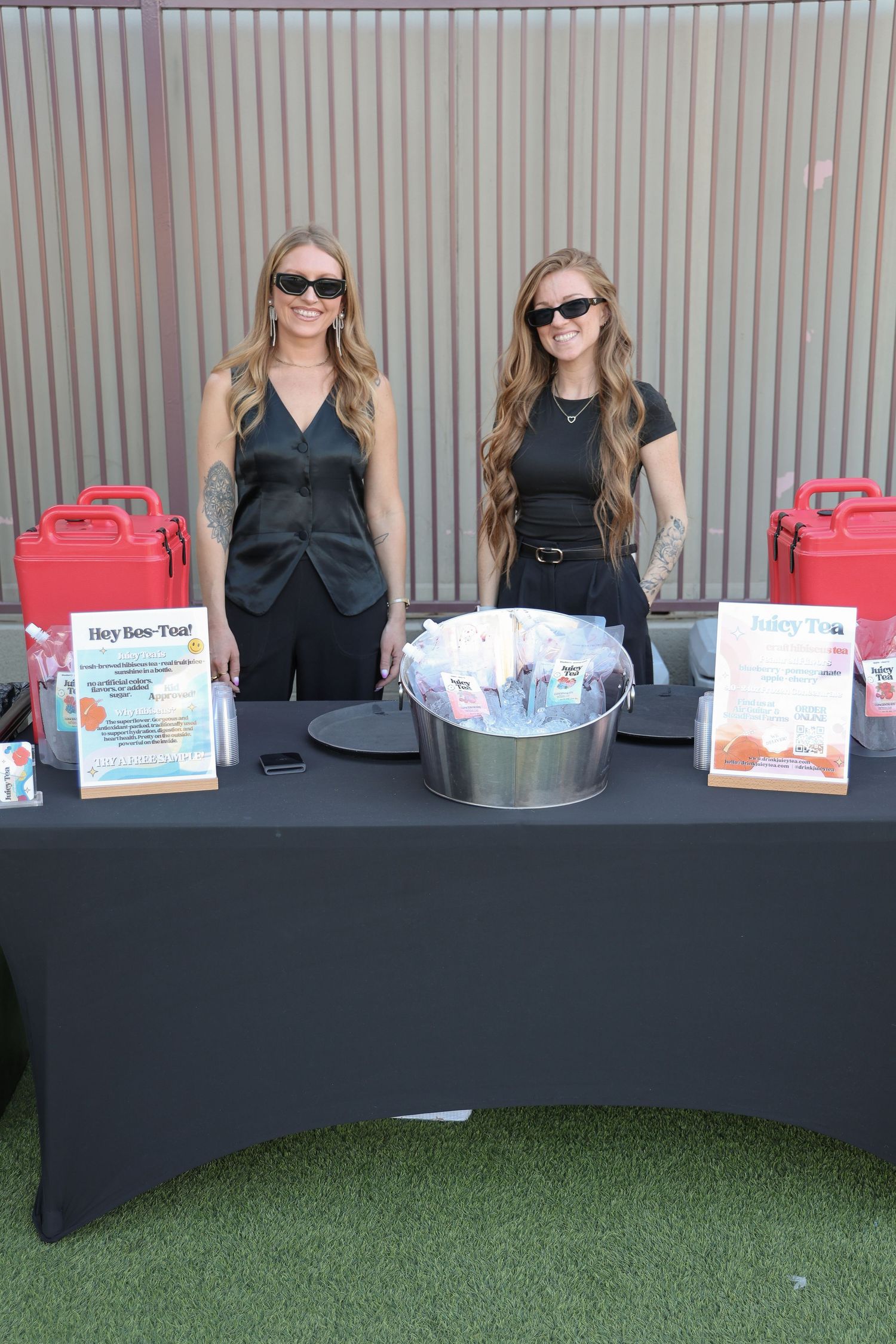 Event booth setup with black tablecloth displaying signs and refreshments on artificial turf.