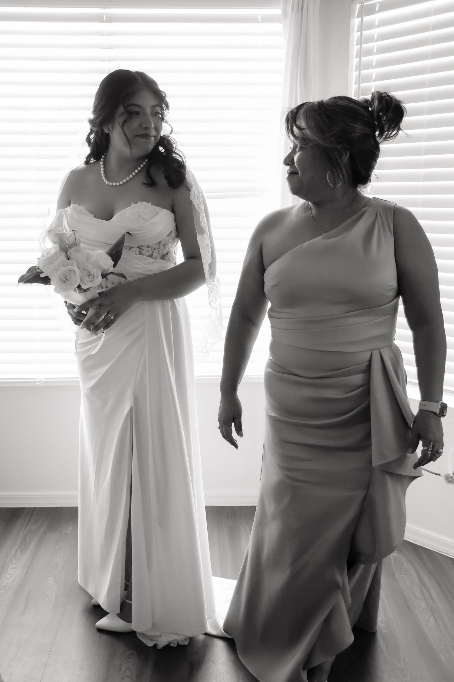 Black and white photo of a bride in white gown holding bouquet standing with a woman in elegant dress near bright windows.