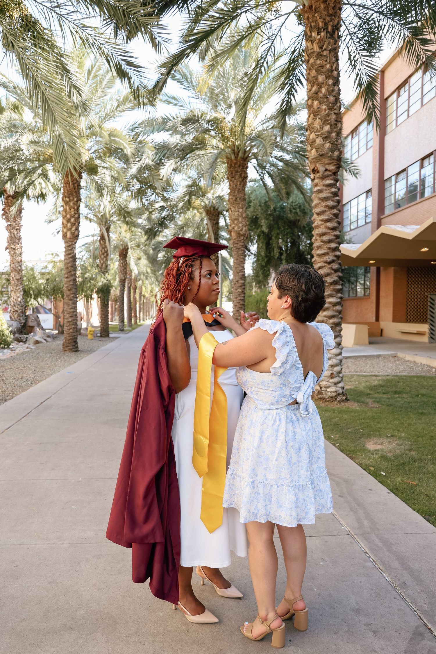 A graduate in maroon cap and gown gets her yellow stole adjusted by a woman in a blue dress near palm trees.