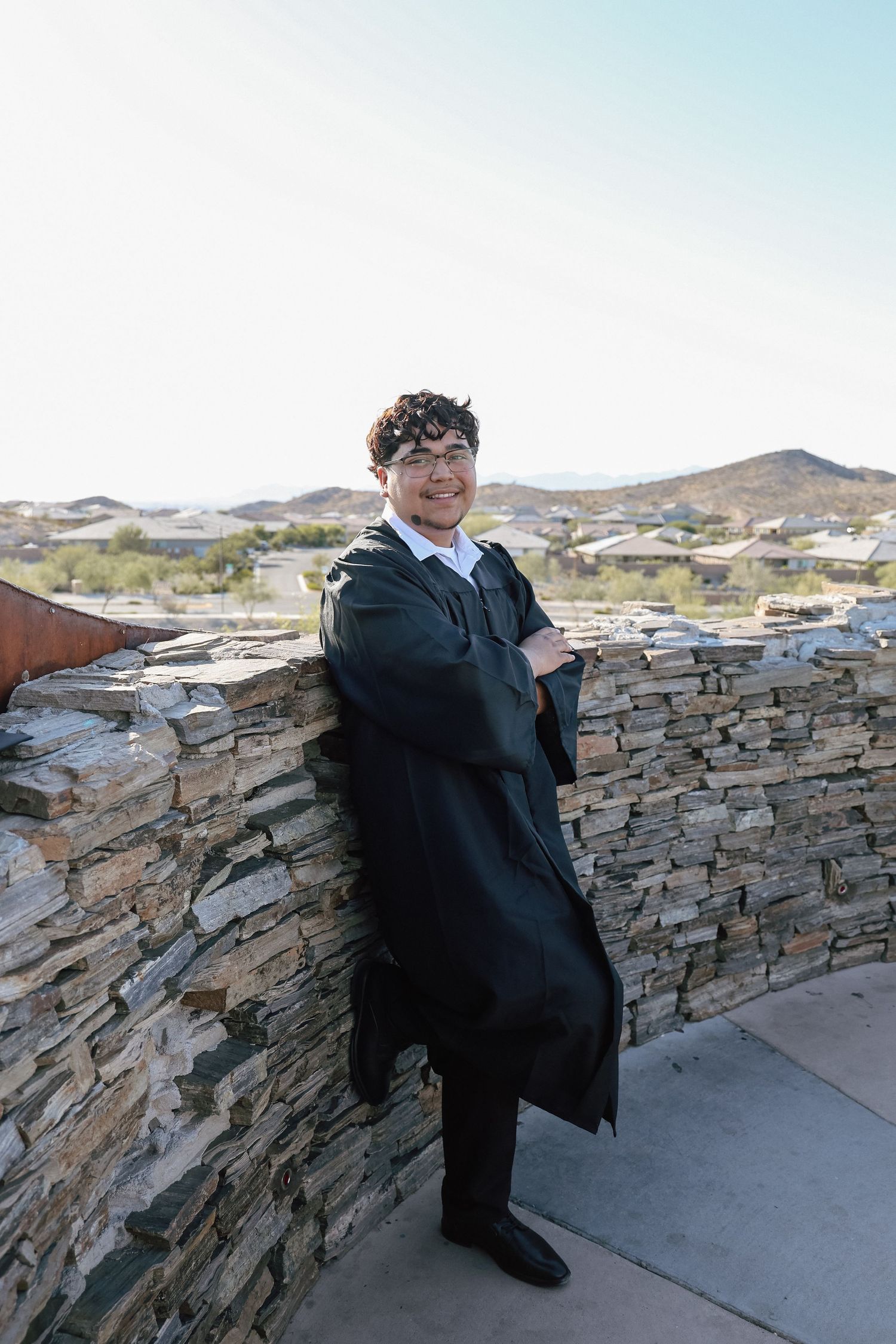 Smiling graduate in black gown leaning against stone wall with desert hills in background.