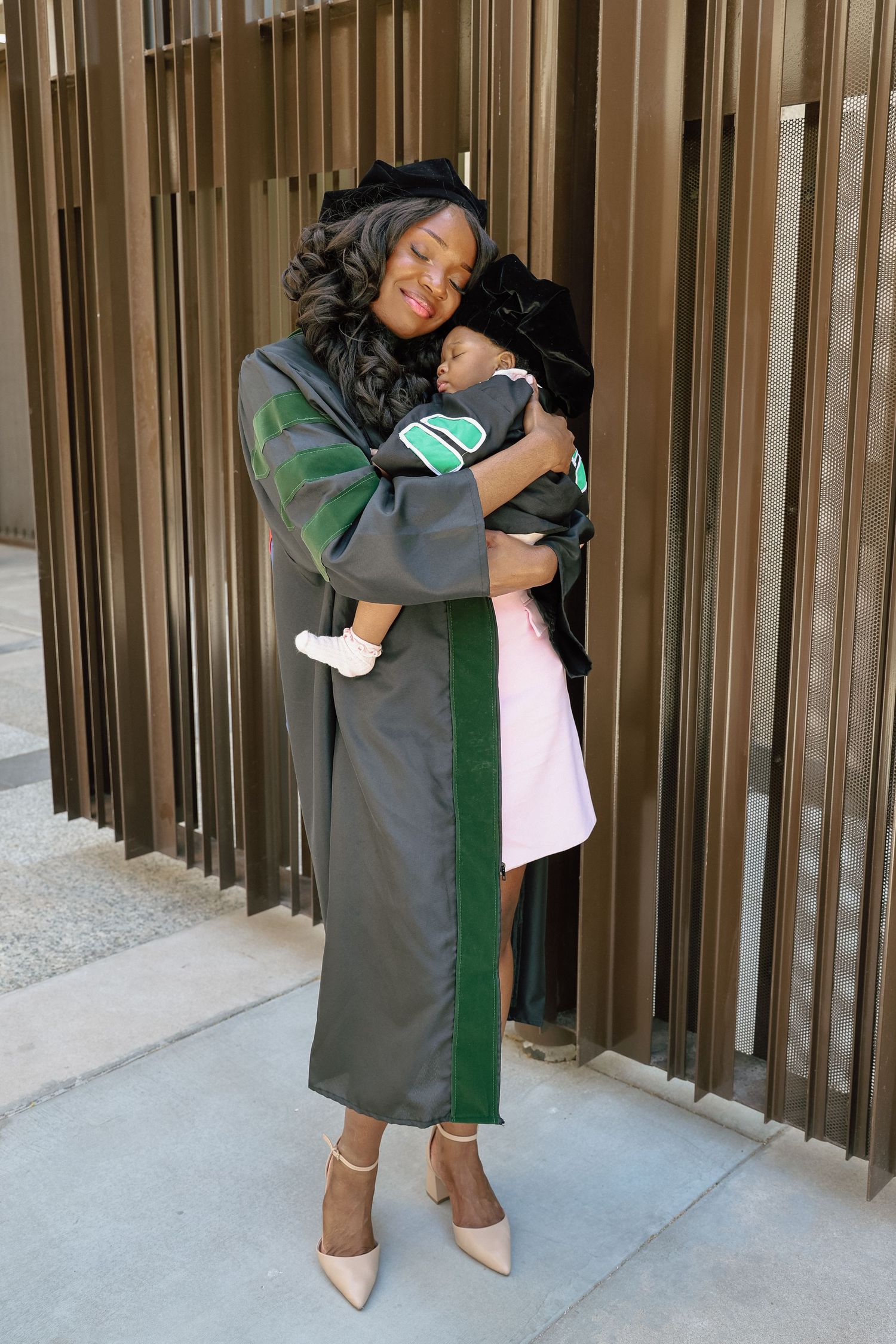 A graduate in green and black academic regalia holds a sleeping baby while smiling outdoors at a graduation ceremony.