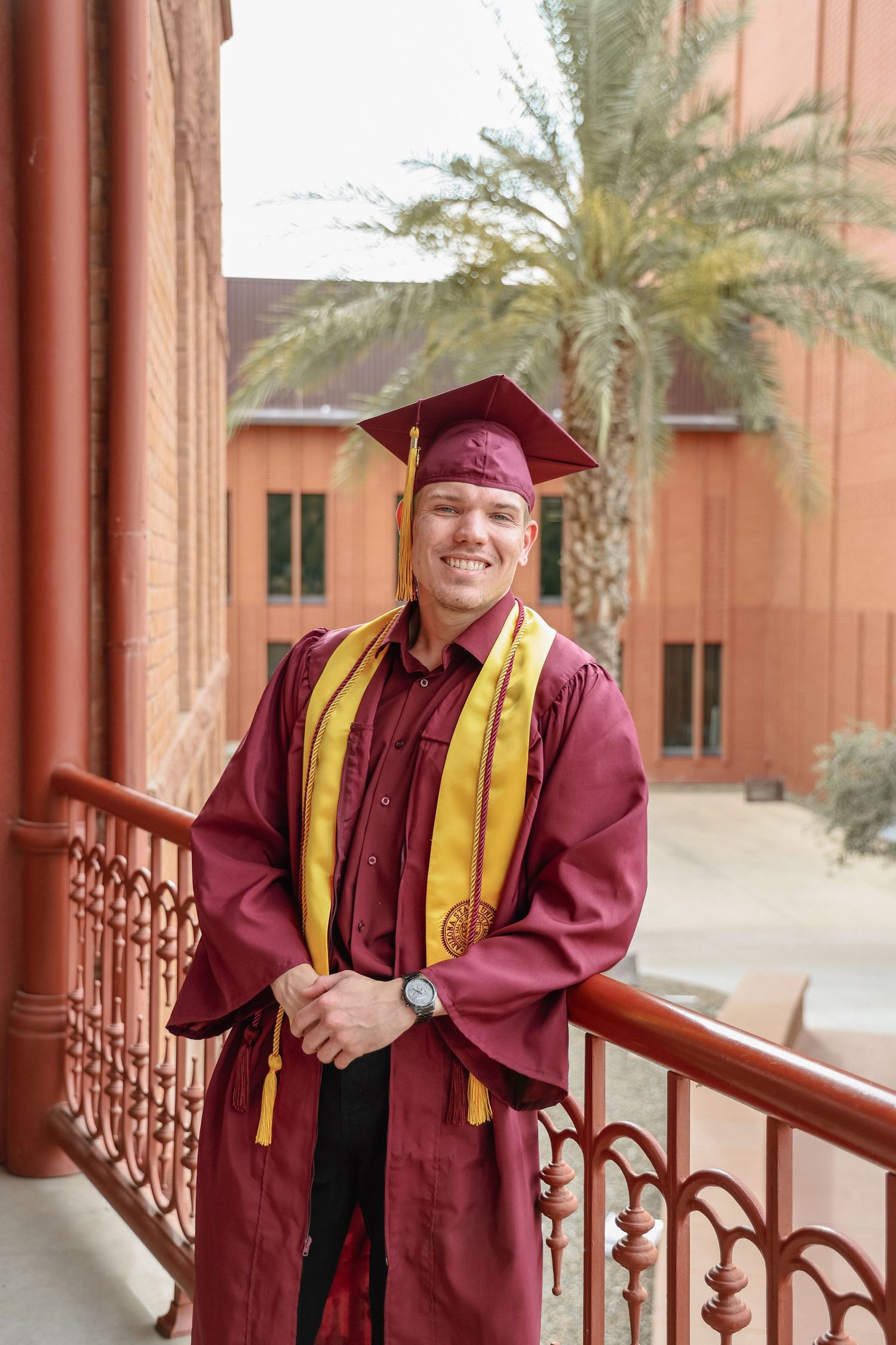 Smiling male graduate in maroon and gold cap and gown stands on a balcony with palm trees and brick building behind him.