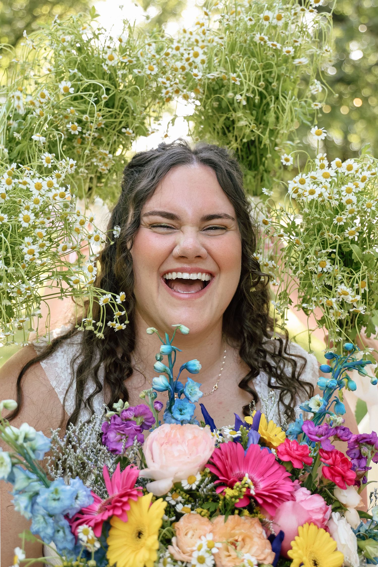 Joyful bride laughing while holding colorful bouquet surrounded by daisy floral arch outdoors.