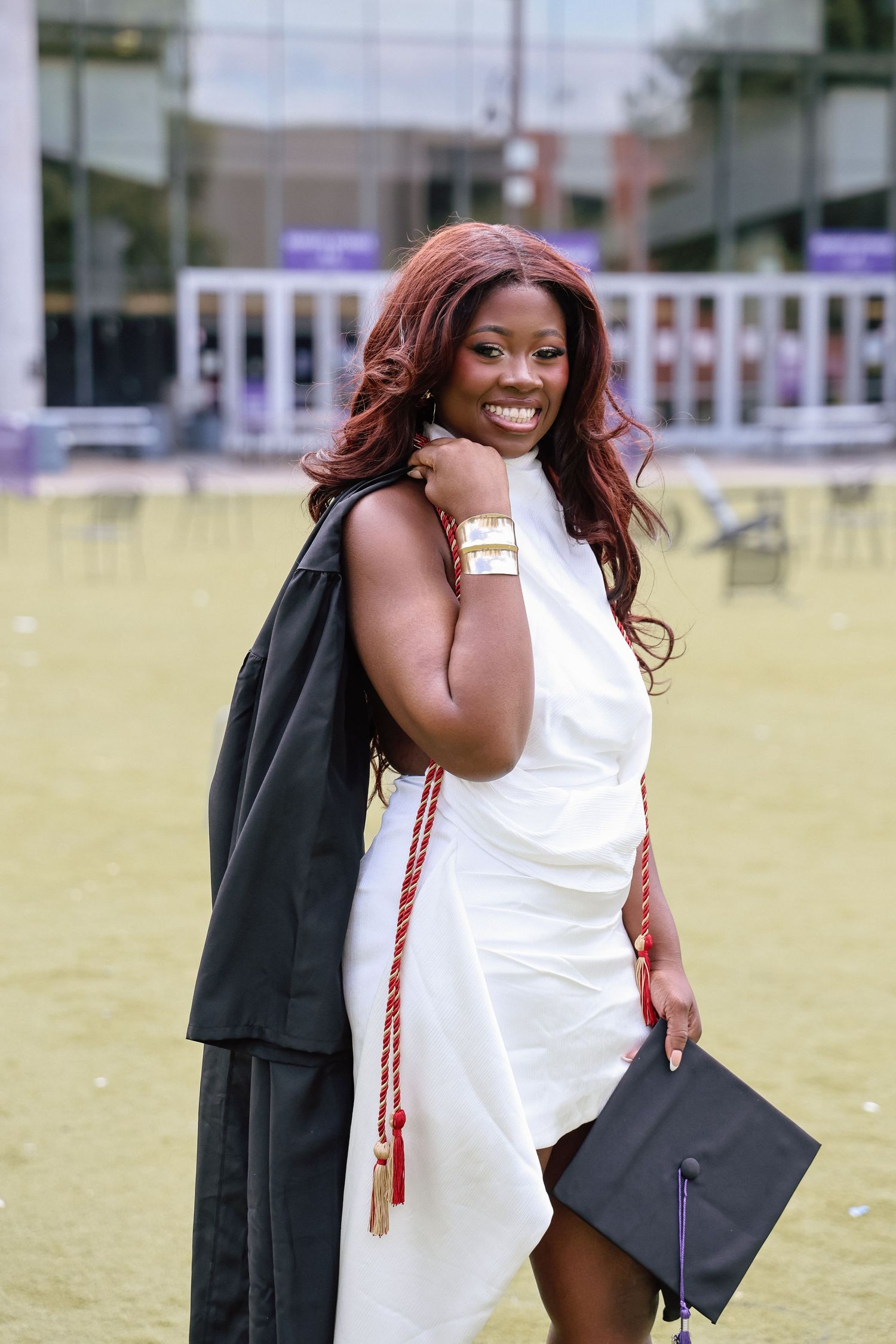 Smiling graduate in white dress holds cap and black gown with red honor cords outdoors on campus lawn.