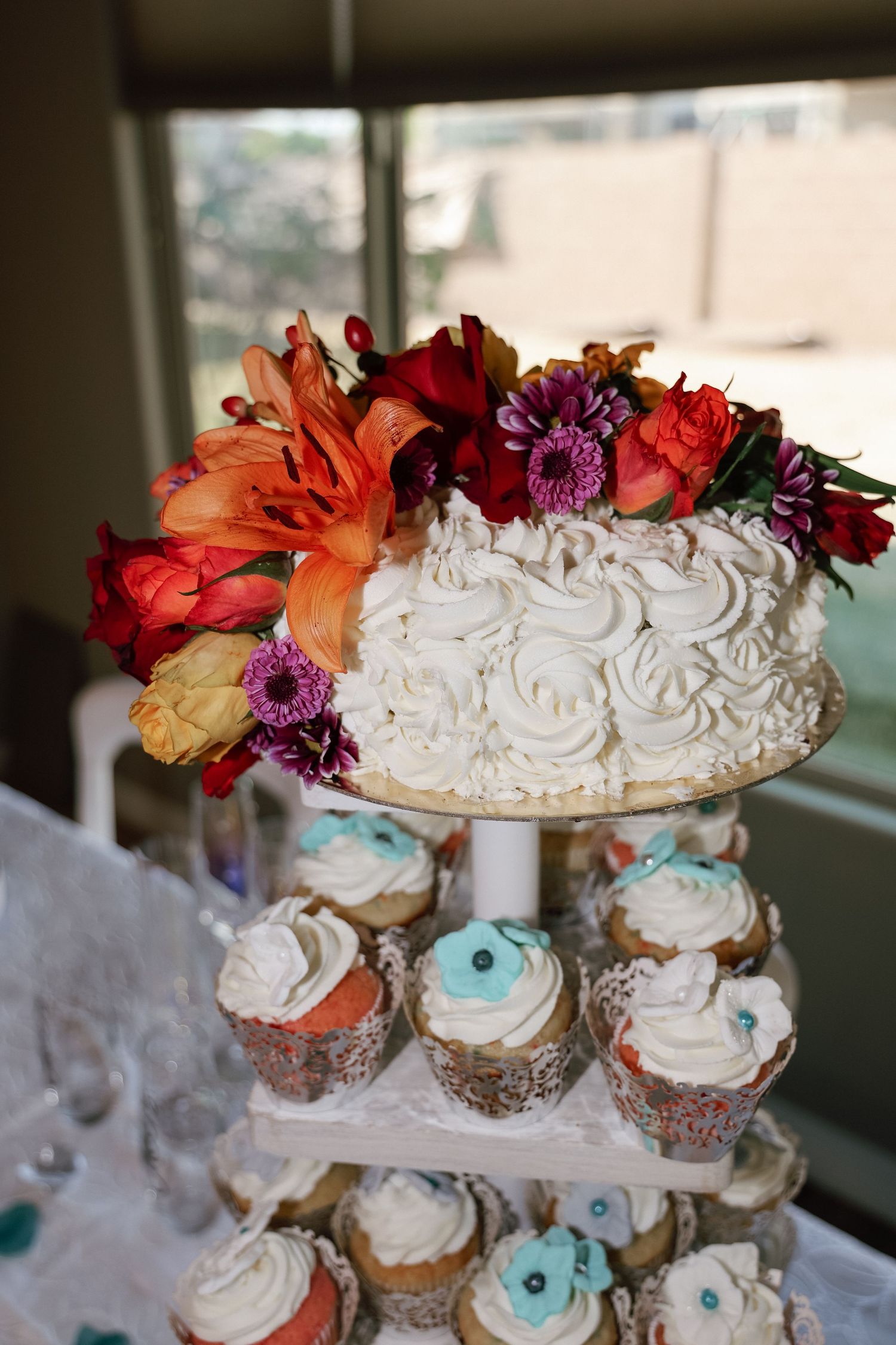 White rosette wedding cake topped with colorful flowers on a tiered stand with decorated cupcakes below.