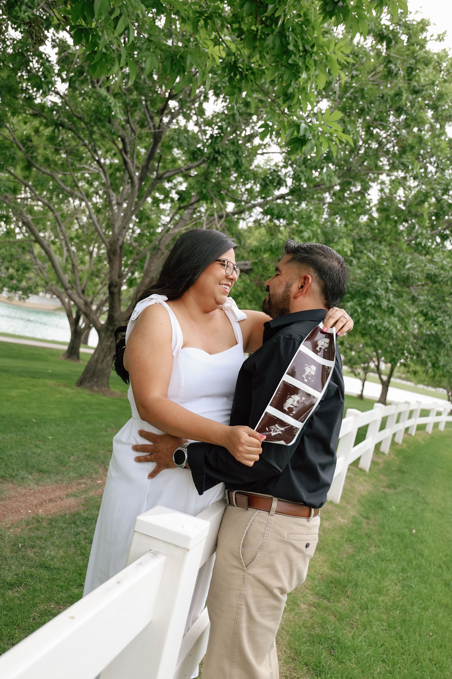 A couple embraces outdoors by a white fence, smiling at each other while holding ultrasound photos, announcing their pregnancy.