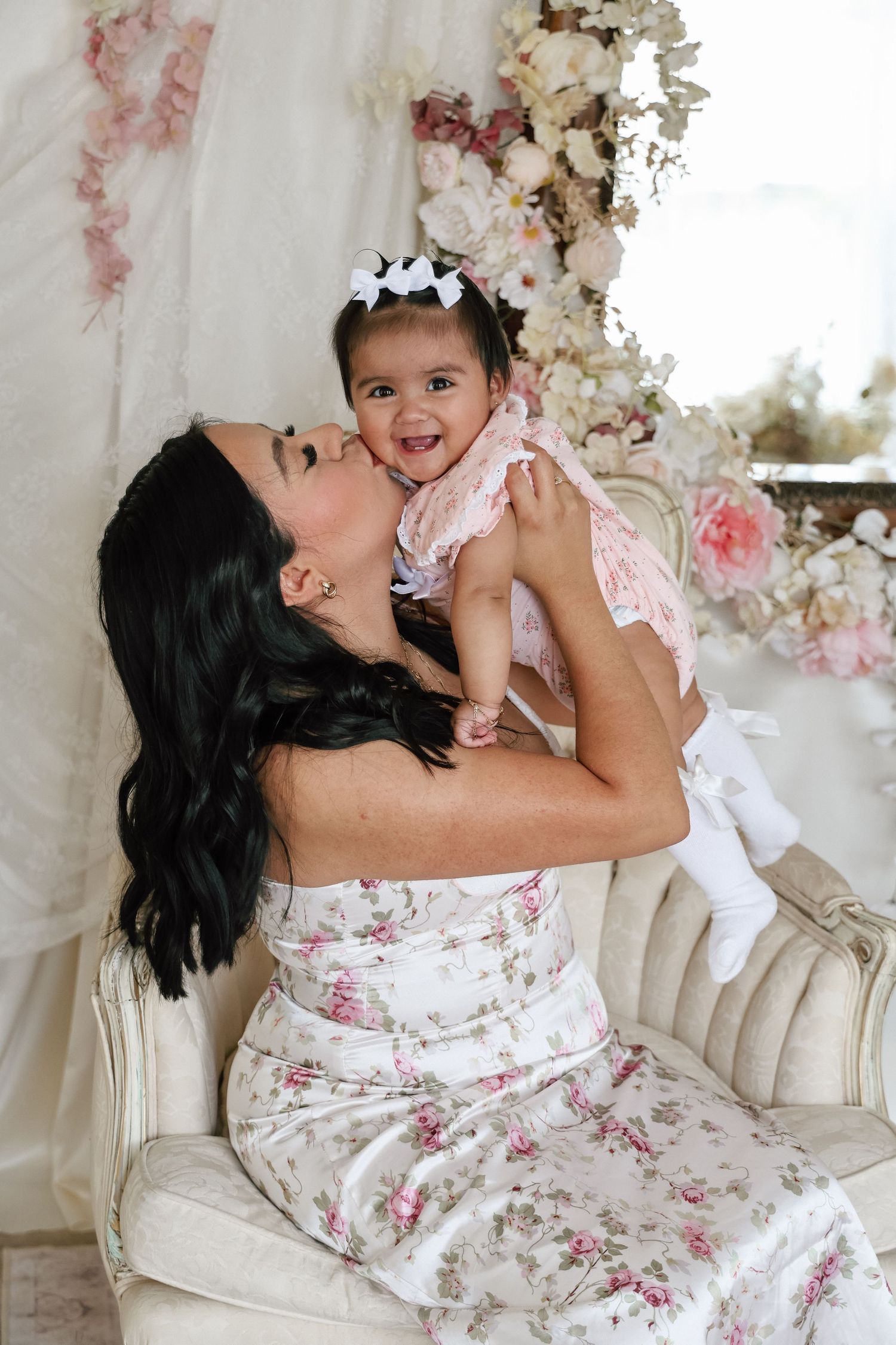 A woman in a floral dress holds a smiling baby girl in pink on a vintage sofa with flower decor.