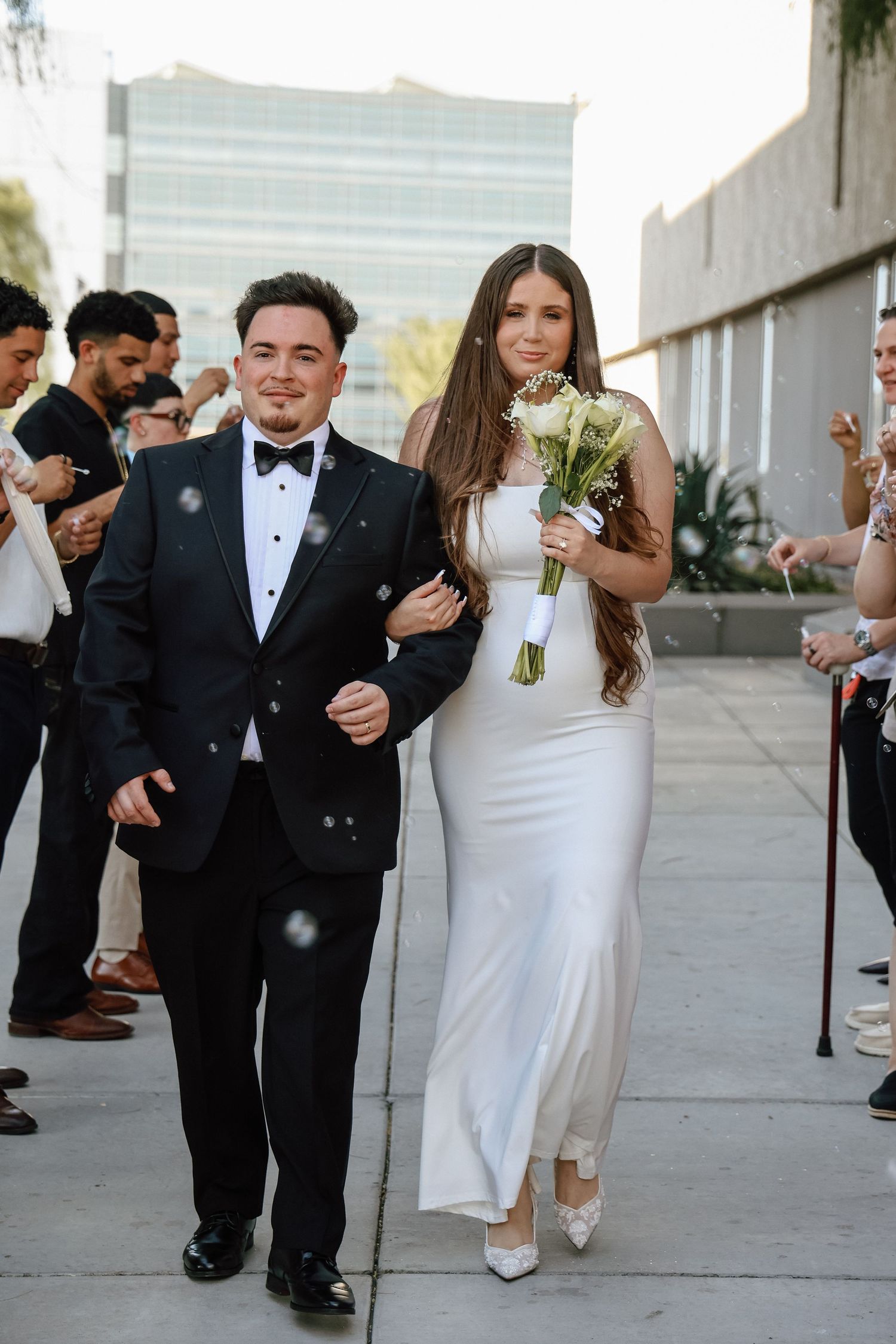 Bride in white gown holding bouquet walks with groom in black tuxedo after wedding ceremony outdoors.
