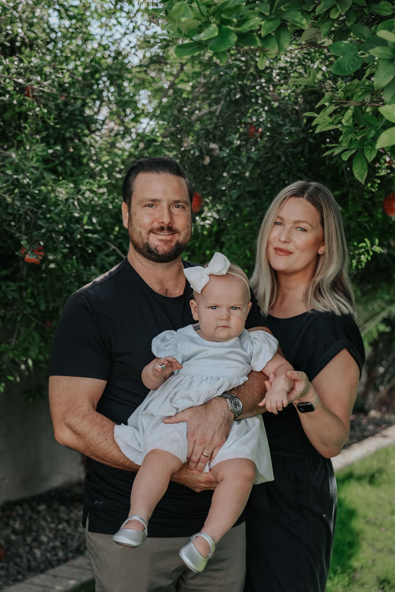 Family of three poses together in front of lush garden backdrop during summer photoshoot.