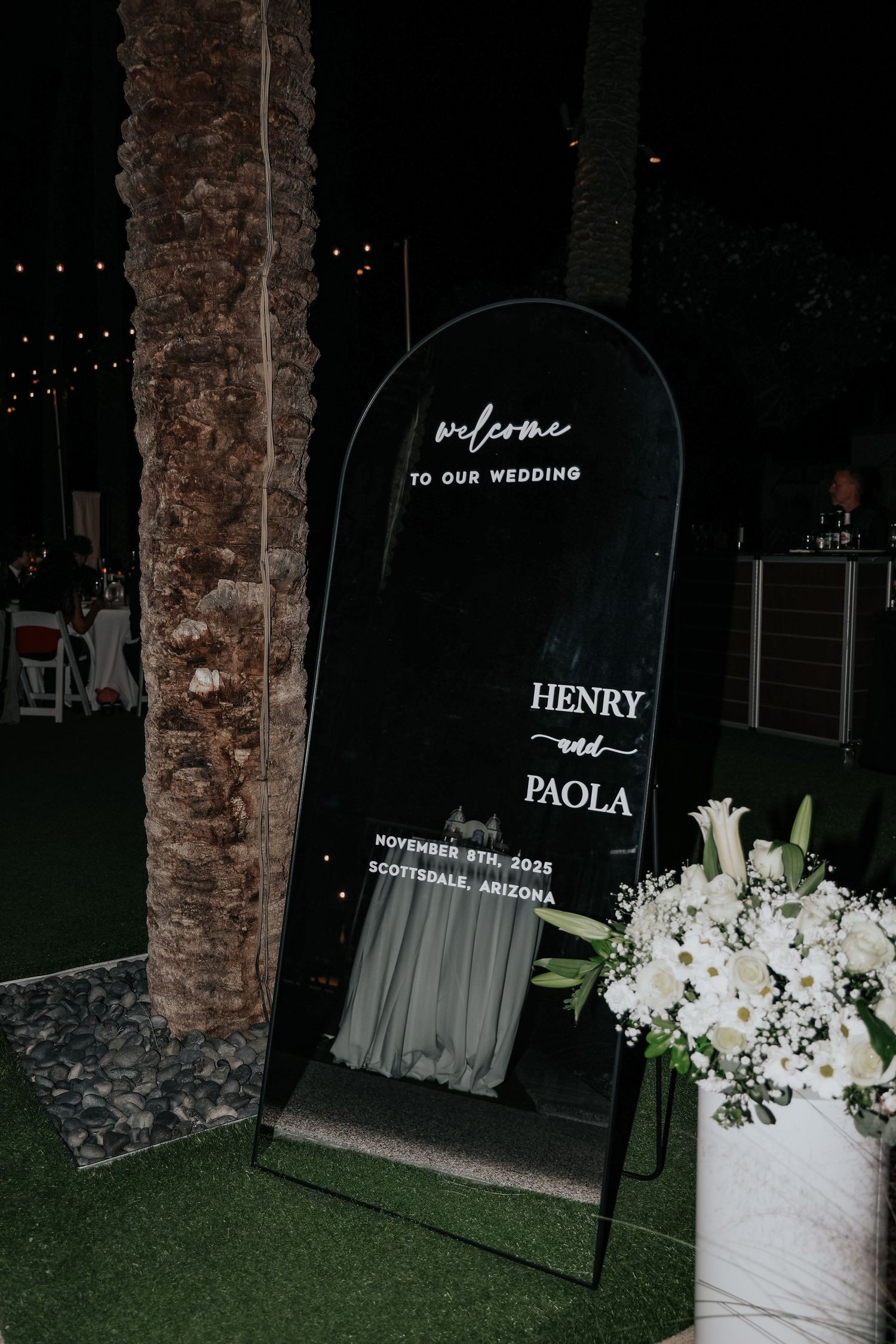 Wedding welcome sign with floral arrangements and neon lighting at night.