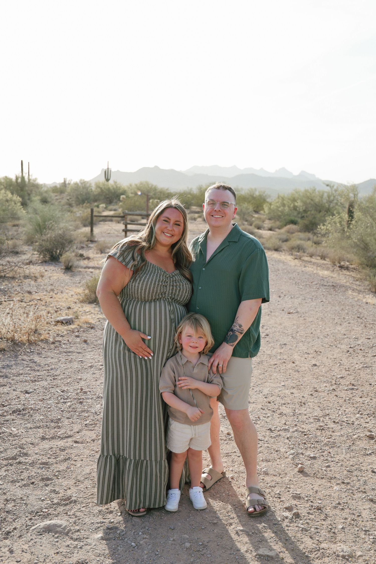 Pregnant woman in green dress poses with husband and toddler son in Arizona desert landscape.