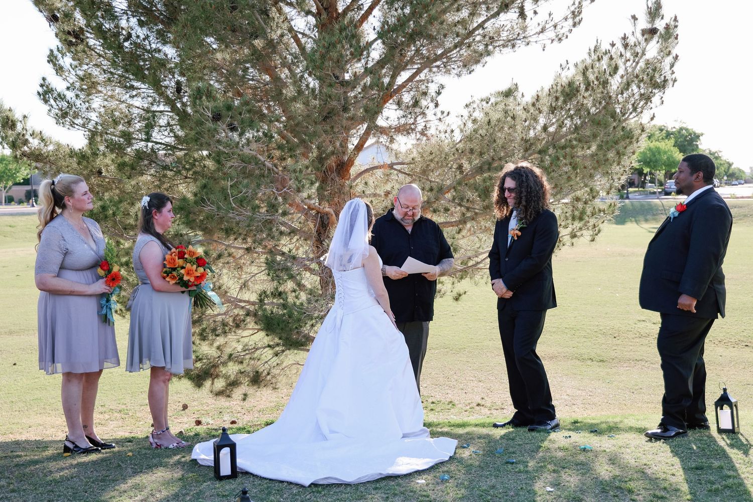 Outdoor wedding ceremony with bride, groom, officiant, bridesmaids, and groomsmen standing on green grass.