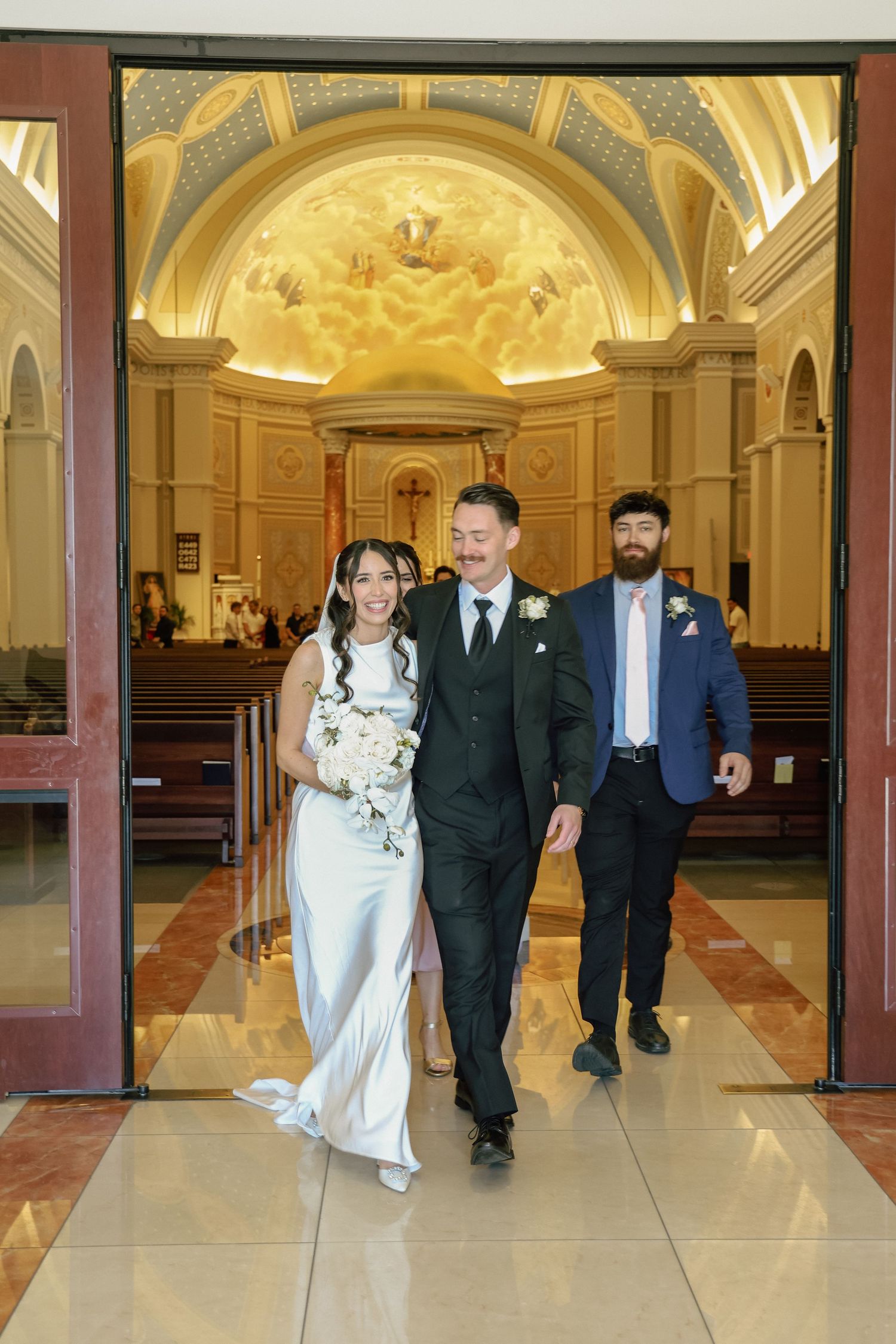 Bride and groom exit a stunning church after wedding ceremony, with bearded groomsman following behind.