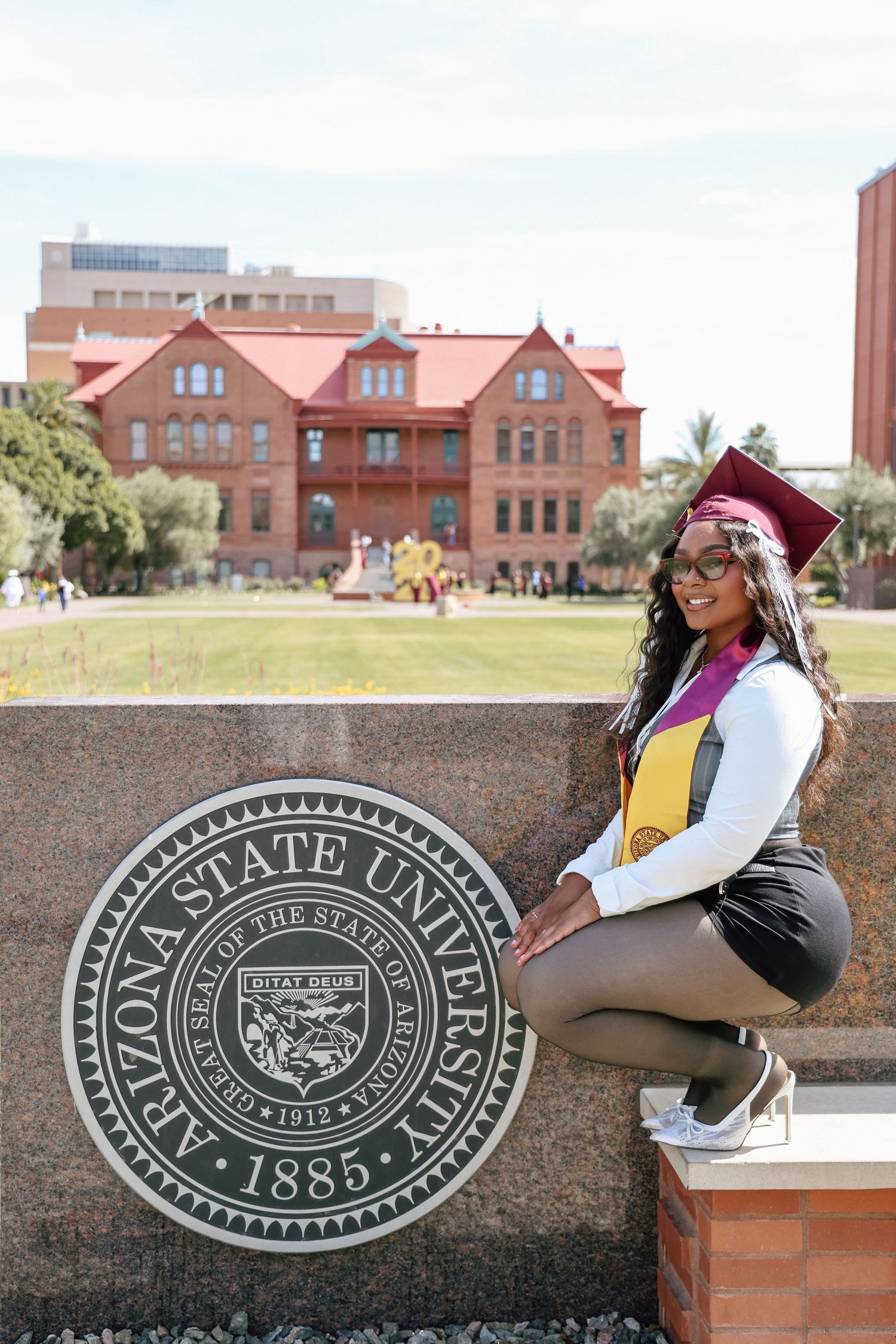 A smiling graduate in cap and gown poses beside the Arizona State University seal on the Tempe campus, 1885.