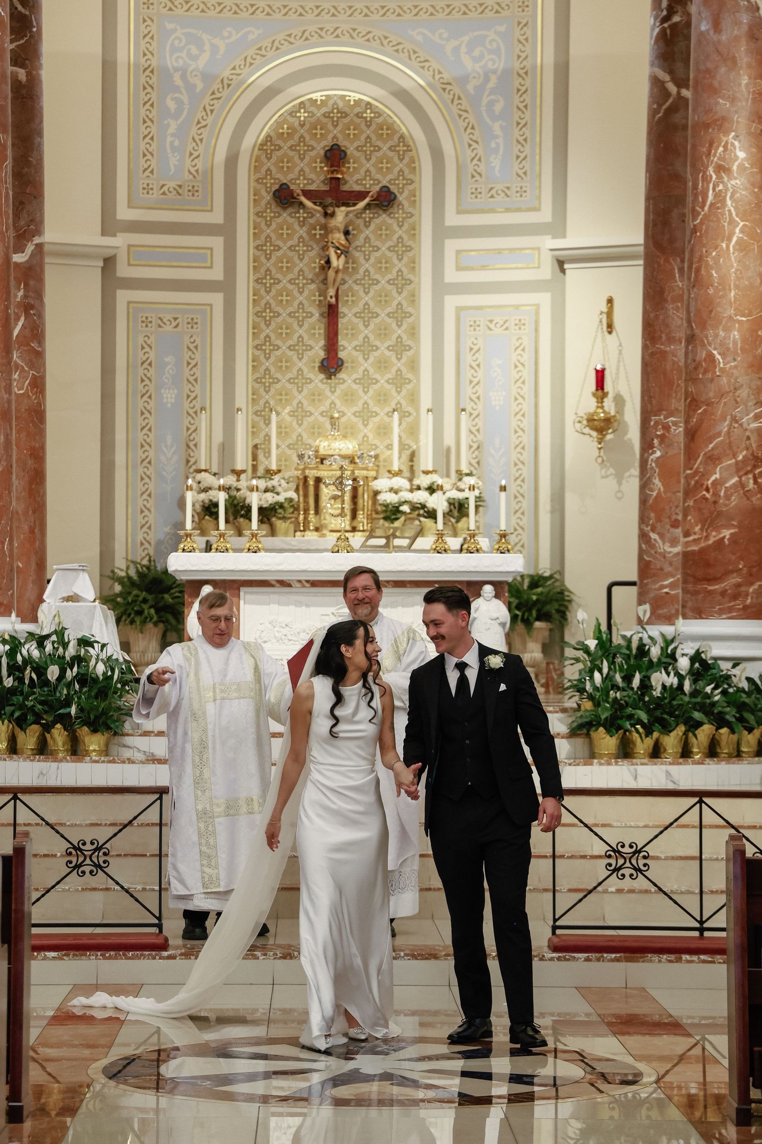 Newlywed couple holding hands at Catholic church altar after wedding ceremony with priest standing behind them.