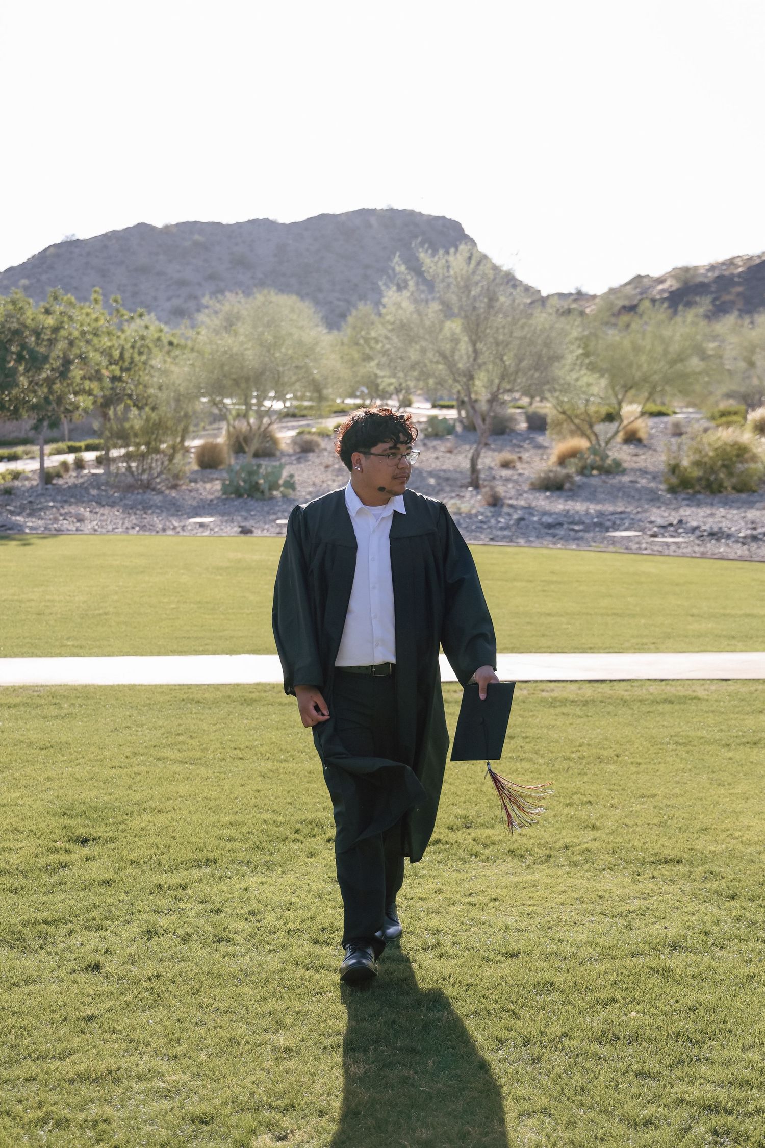 A graduate in green cap and gown walks across a lawn with desert mountains in the background.