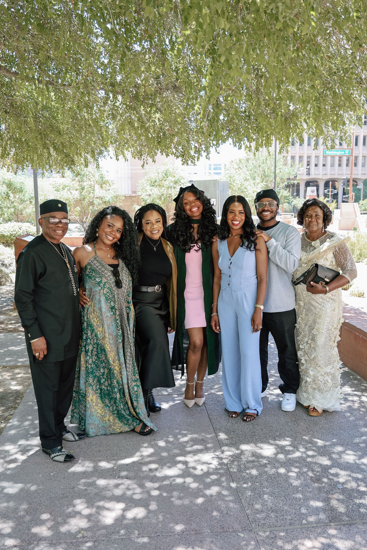 A joyful family group of seven poses outdoors under a tree, dressed in elegant formal attire for a graduation celebration.