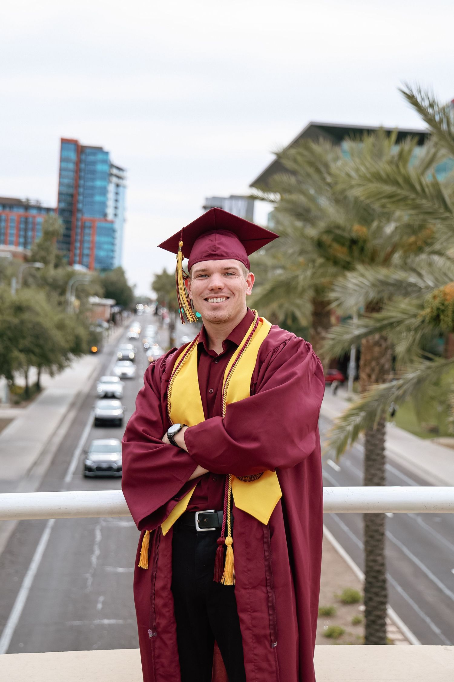 Male graduate in maroon cap and gown poses with arms crossed on an urban bridge overlooking a palm-lined street.