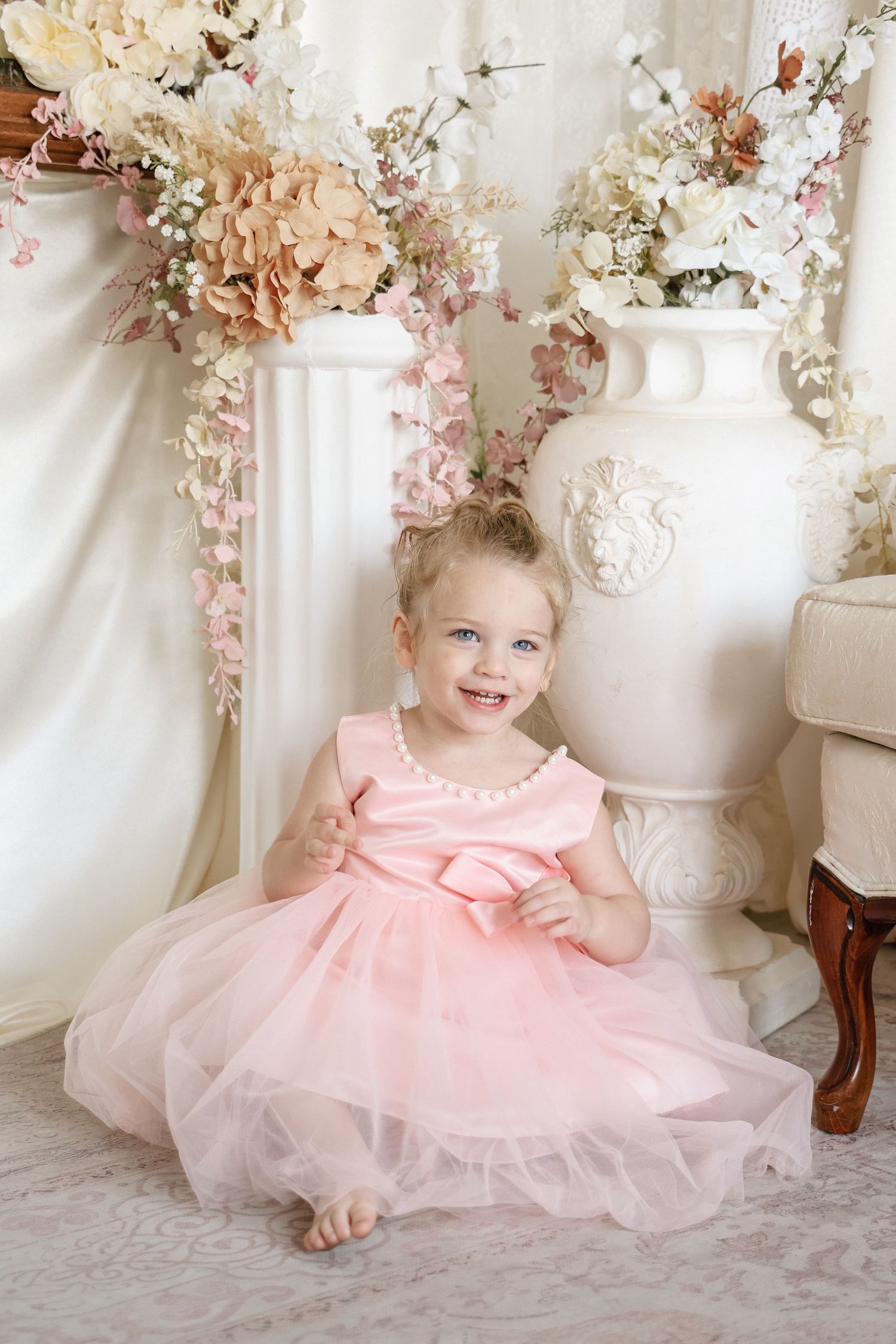 Smiling toddler girl in pink tulle dress with pearl neckline sits on floor beside white vase with flowers.