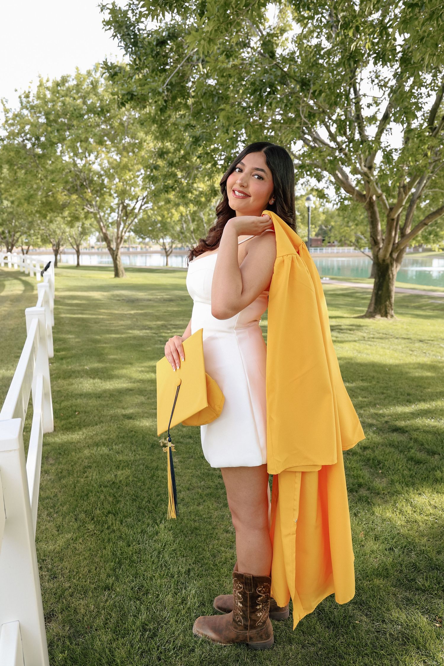 Smiling graduate in white dress poses with yellow cap and gown beside white fence in green park.