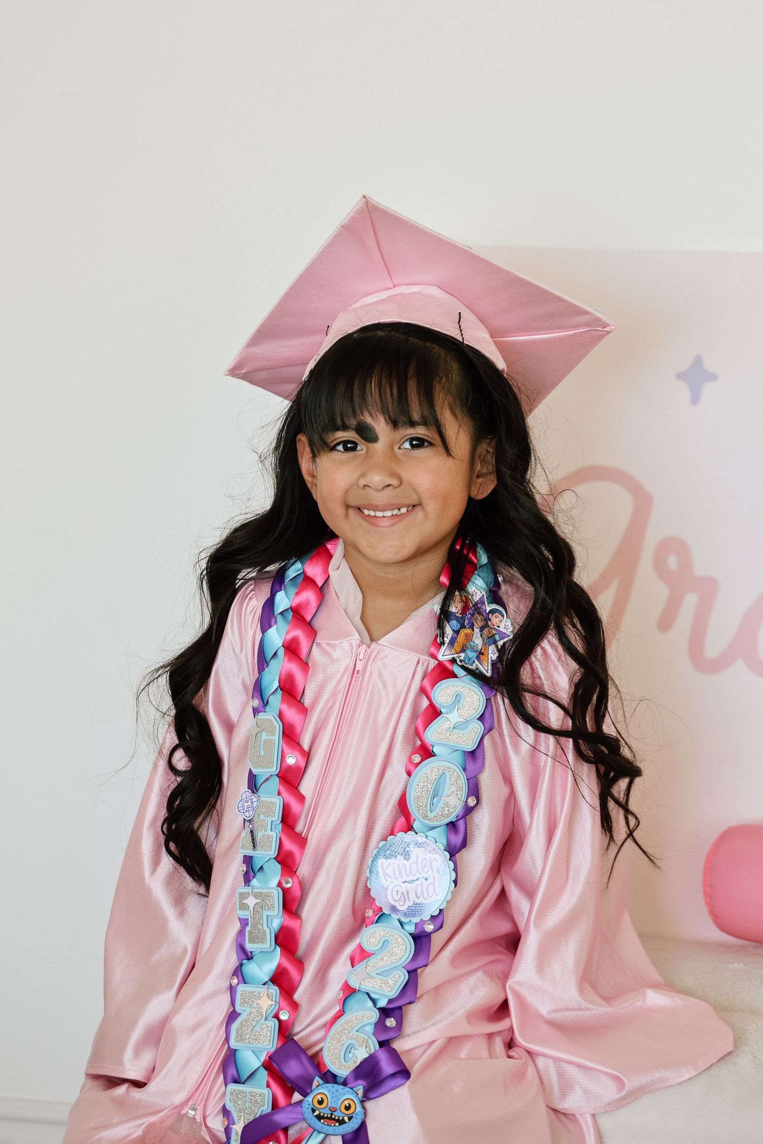 Young girl in pink graduation cap and gown with colorful sash smiling for her preschool graduation photo.
