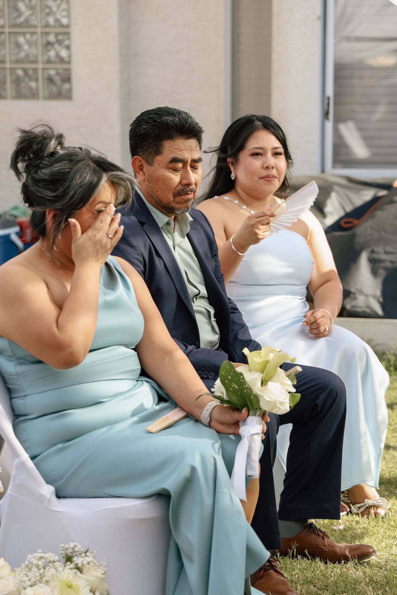 Emotional wedding guests in blue formal attire, with a woman crying and another holding a bouquet during an outdoor ceremony.