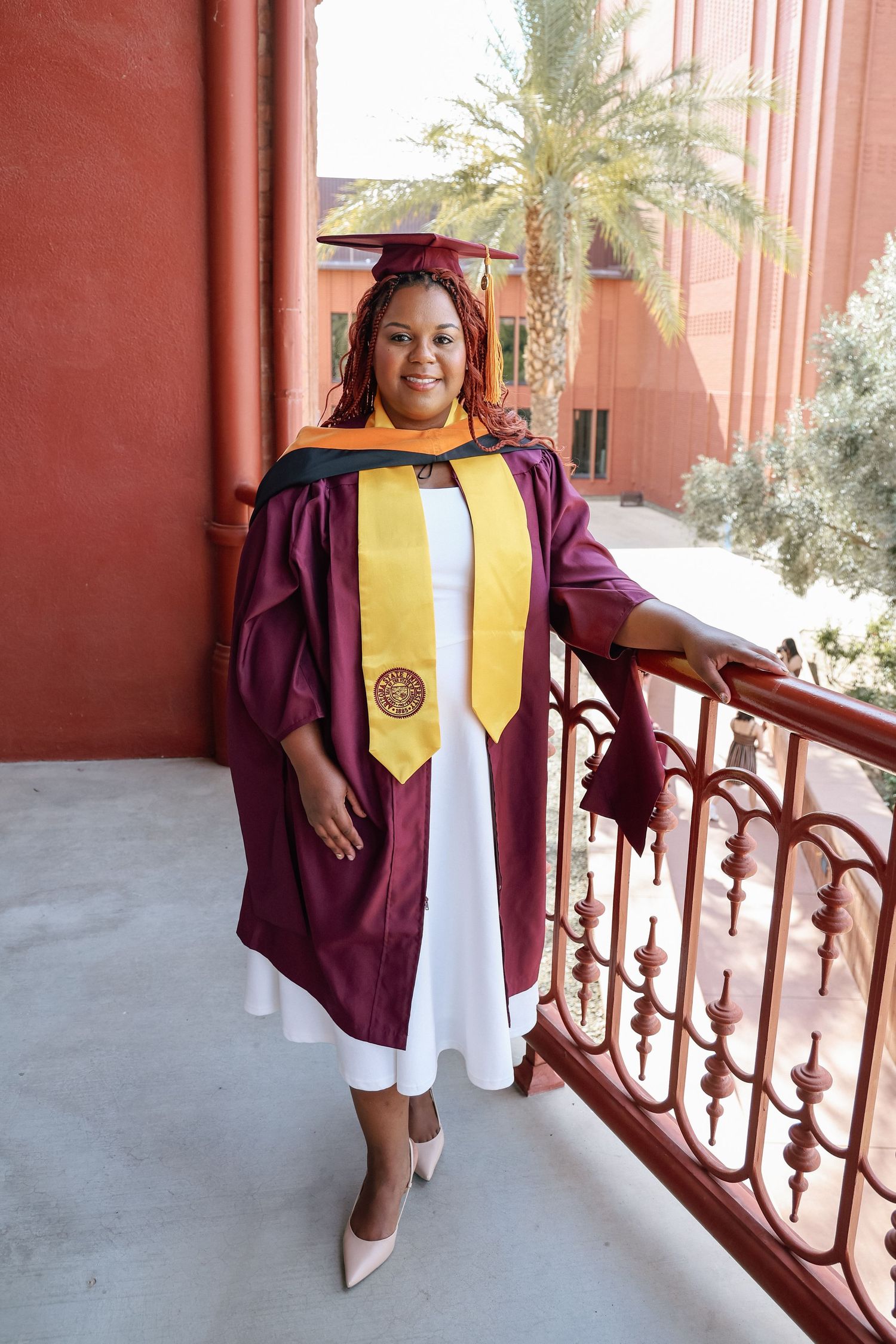 A woman in maroon graduation gown and cap with gold stole poses proudly on a balcony railing outdoors.