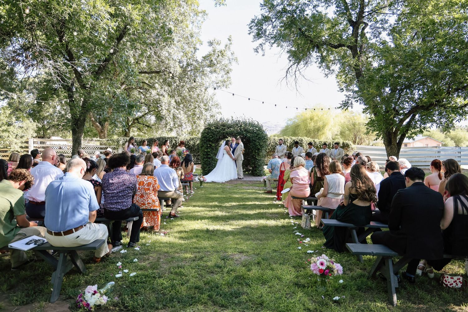 Outdoor wedding ceremony on green lawn with guests seated on benches as couple exchanges vows under floral arch.