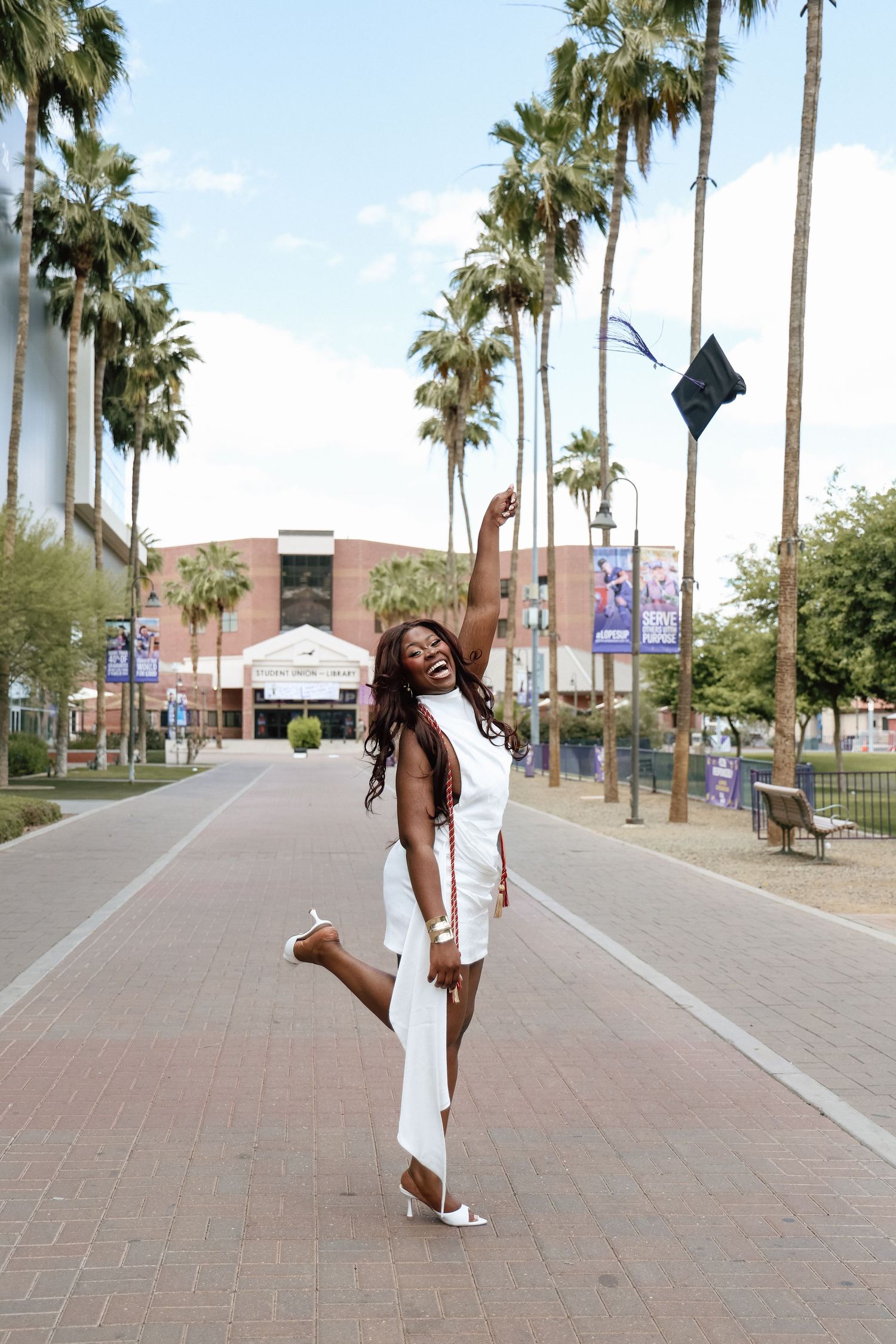 A joyful Black woman in a white dress tosses her graduation cap in the air on a palm-lined campus walkway.