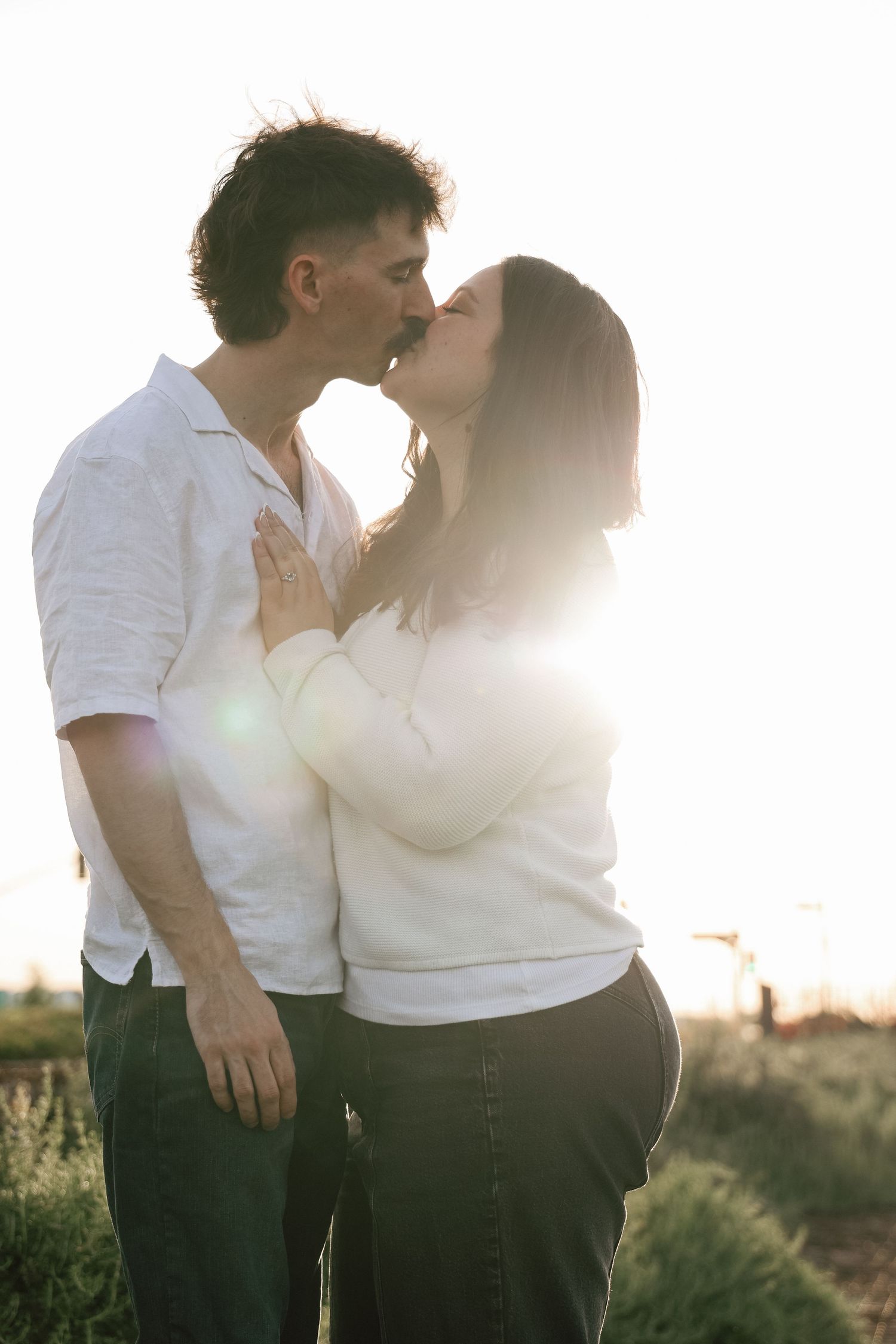 A romantic couple in white shirts share a tender kiss at sunset with sunlight creating a warm backlit glow.