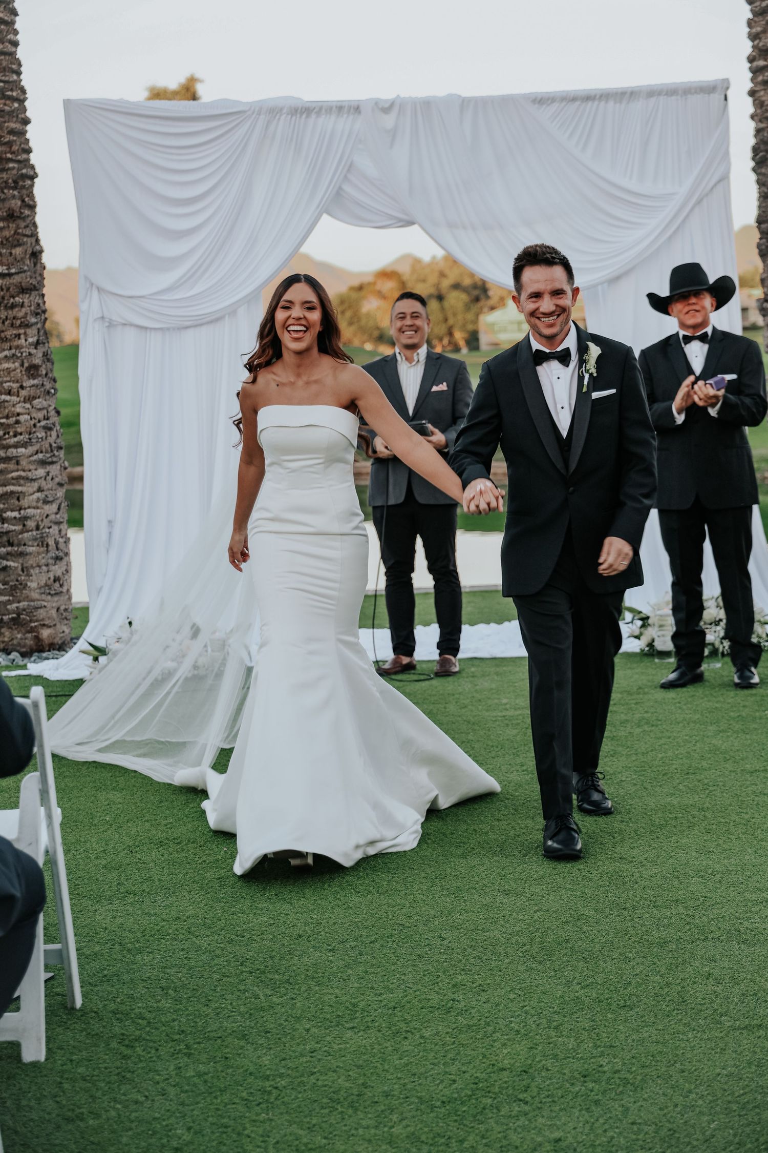 Wedding party running and laughing on a green lawn with white drapes in the background.
