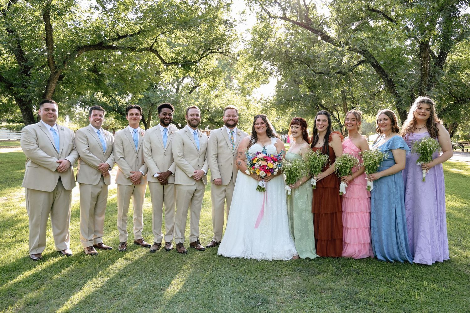 Wedding party poses outdoors in a lush green park, groomsmen in tan suits and bridesmaids in colorful dresses flanking the bride and groom.