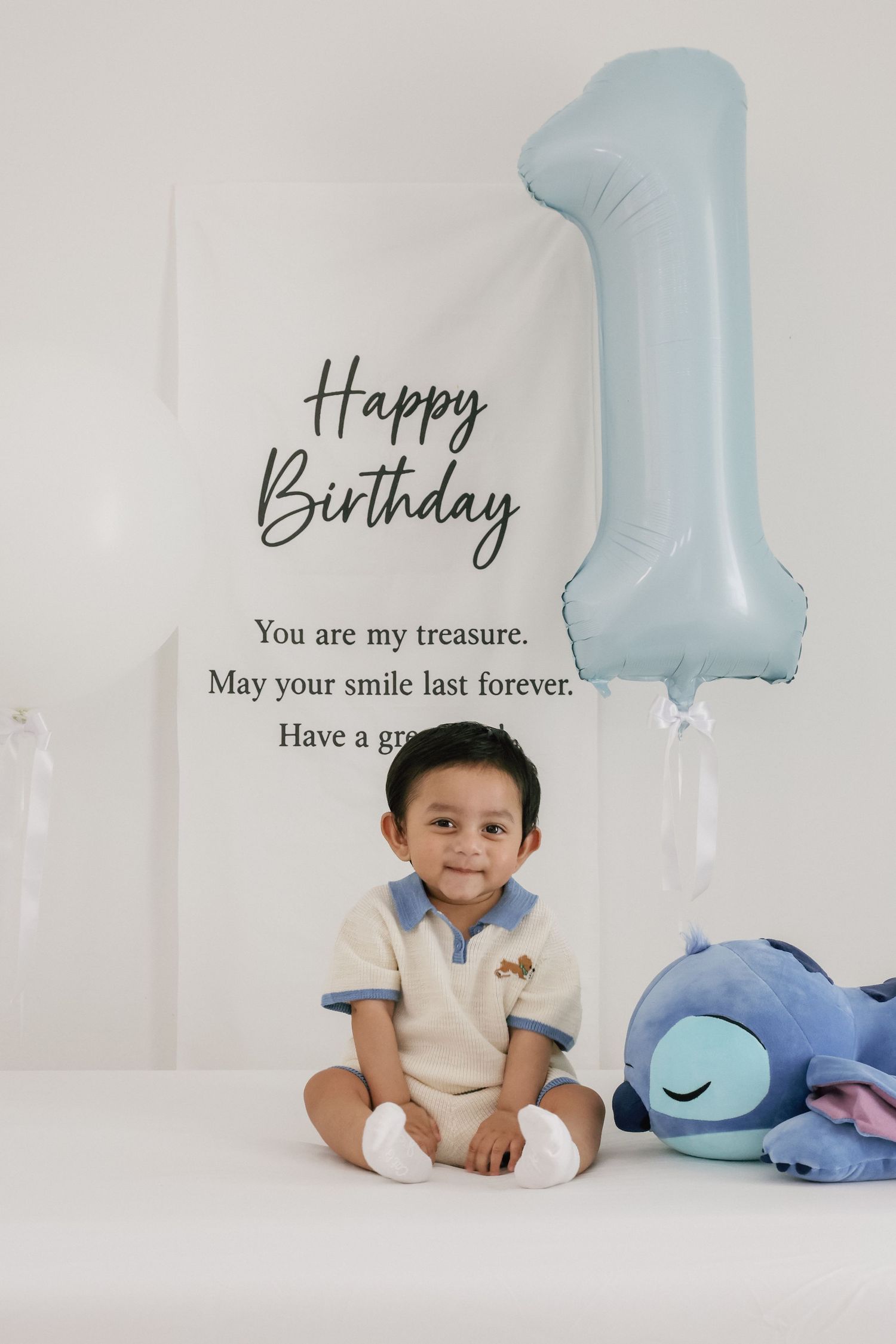 A smiling baby boy celebrates his 1st birthday with a blue number one balloon and Stitch plush toy.