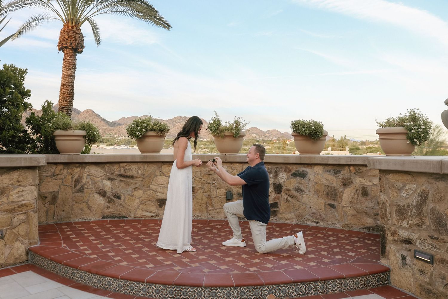Man proposes to woman on one knee at scenic outdoor terrace with palm trees and mountain views.